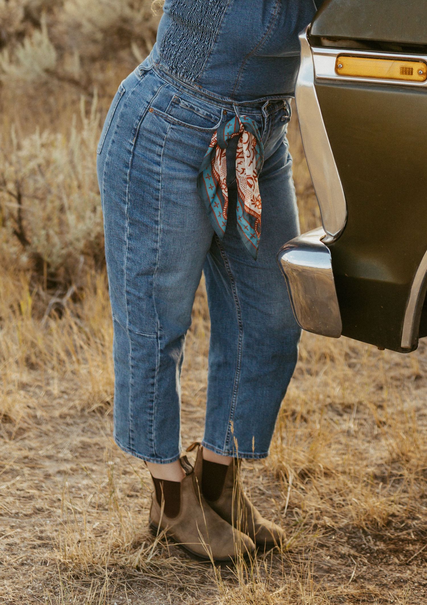 Woman wearing cropped jeans, a bandana belt, and brown ankle boots standing by a vintage car.