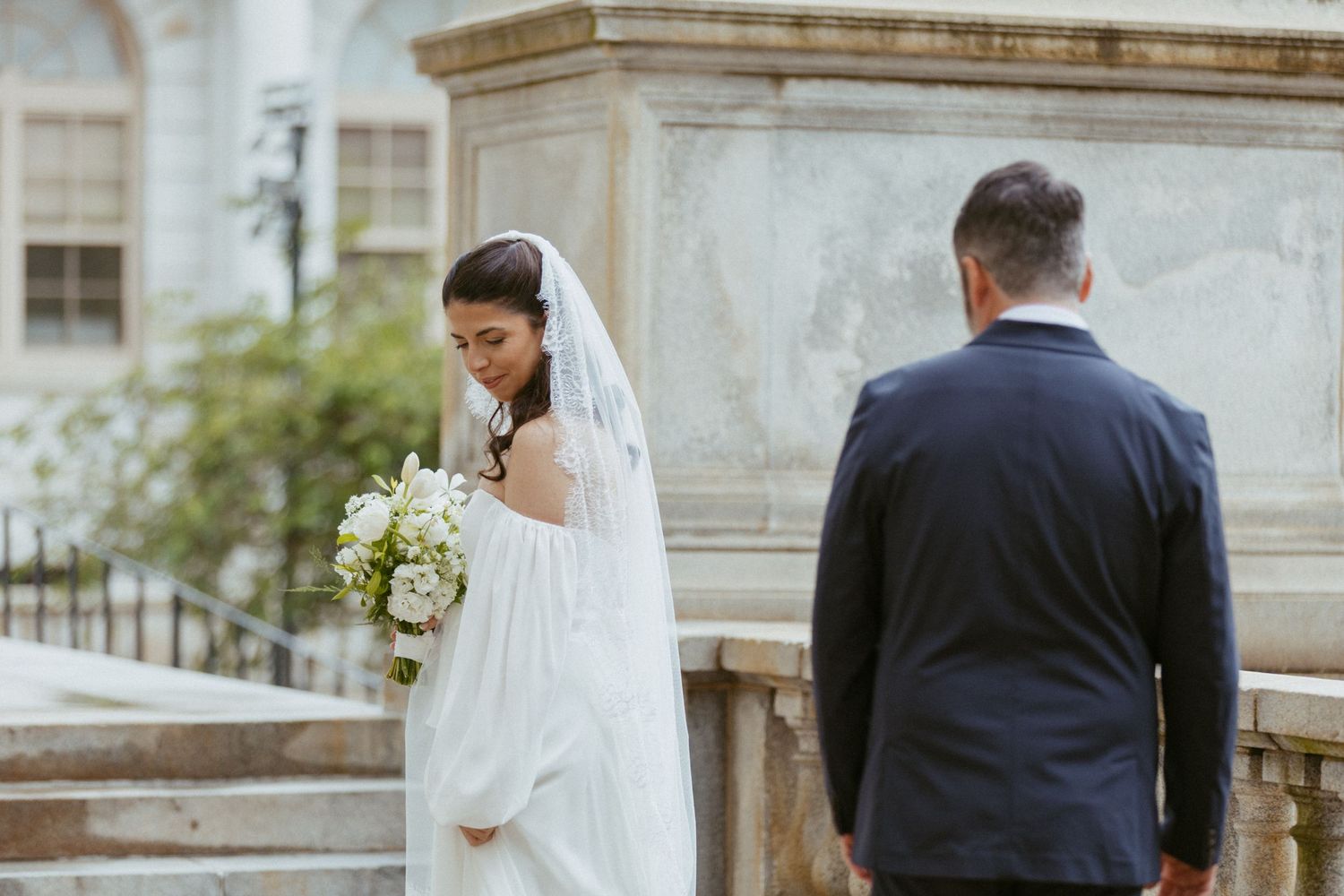 Bride in white gown and veil holds bouquet while groom stands nearby at a stone building.