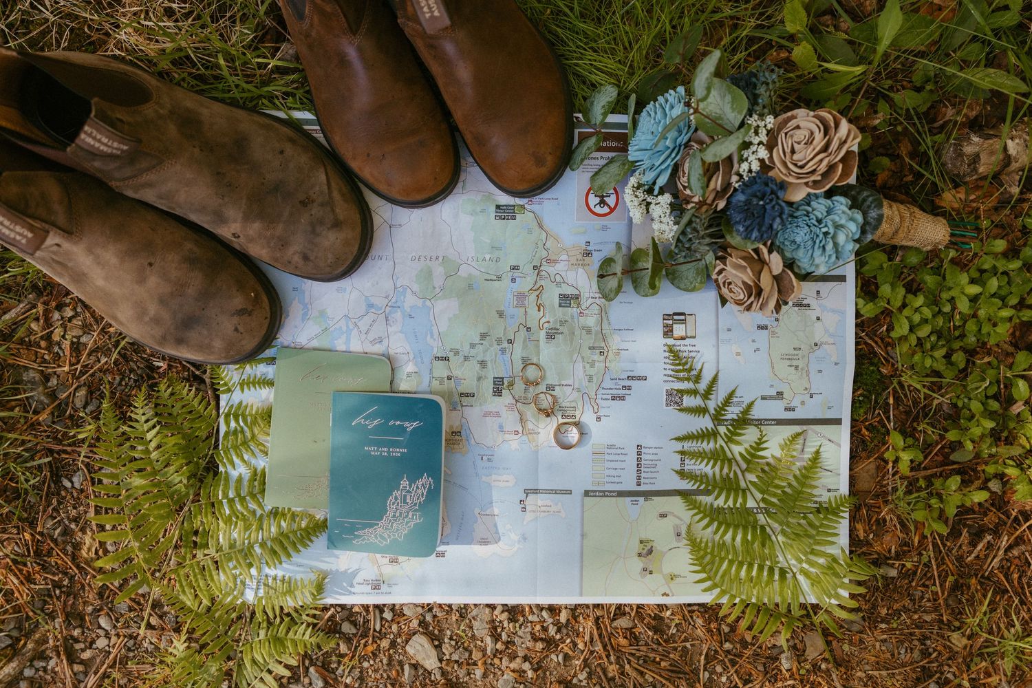 Rustic wedding flat lay with map, blue floral bouquet, leather boots, ferns, and navy vow books on grass.