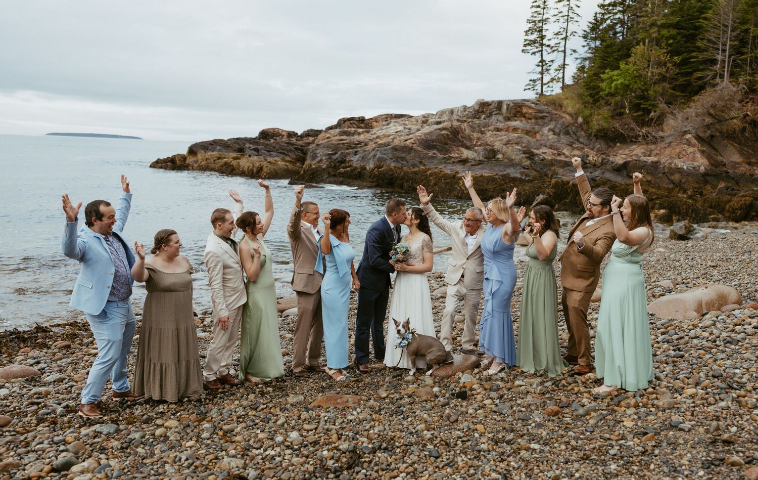 Wedding party celebrating on rocky beach with ocean backdrop, bridesmaids in sage and blue gowns, arms raised in joy.