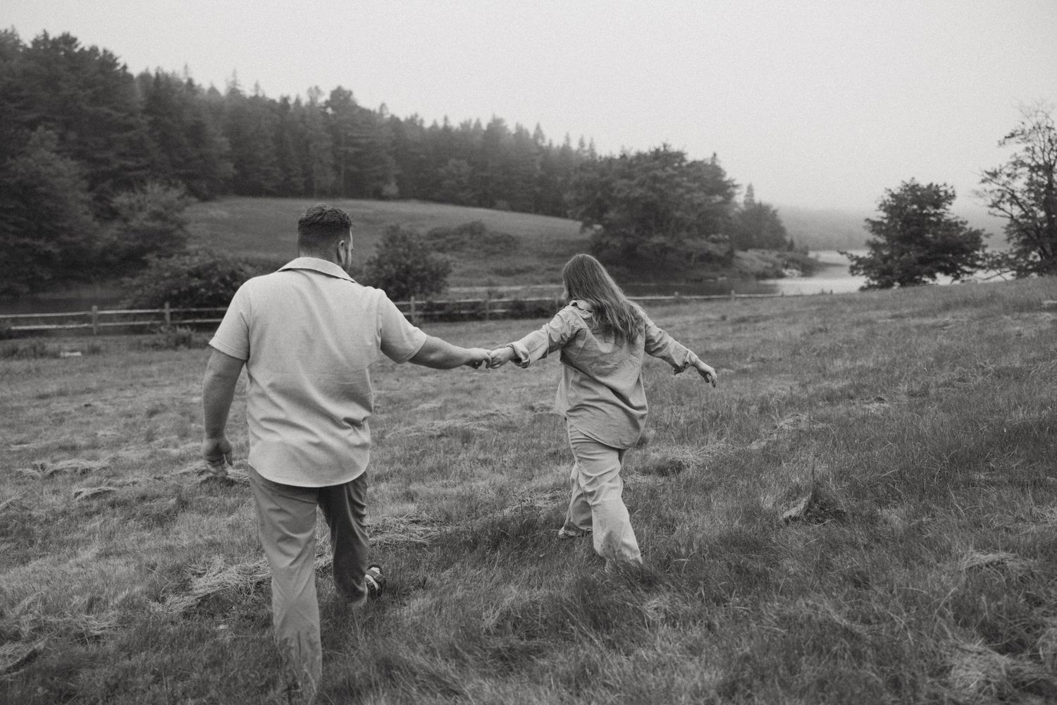 Black and white photo of a couple holding hands while walking through a grassy field with trees in the background.