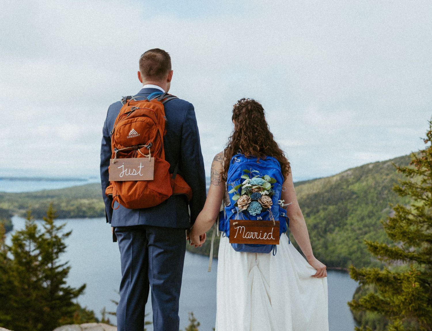 Newlywed couple holding hands with Just Married backpacks at scenic overlook in wedding attire.
