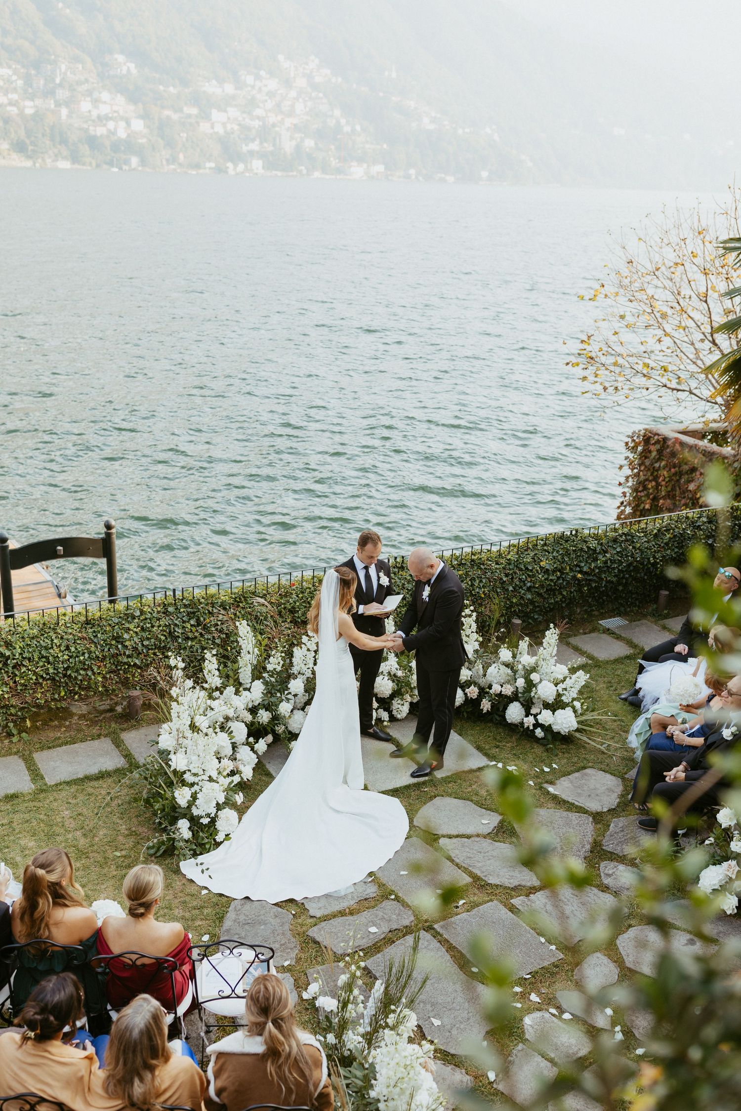 Bride and groom hold hands at an outdoor lakeside wedding ceremony with stunning water views and white floral arrangements.