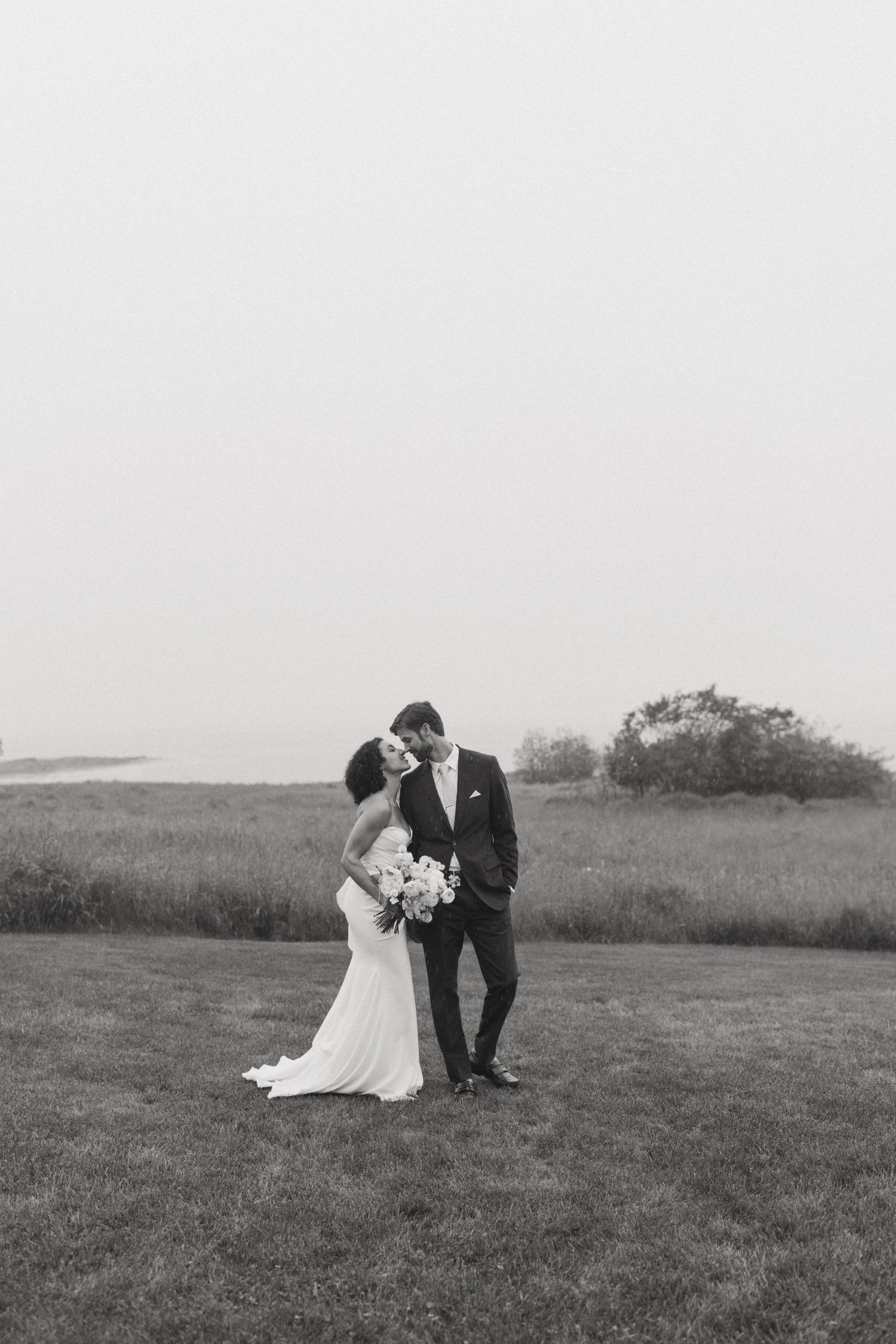 Bride and groom kissing on grassy lawn with ocean view in black and white wedding photography.