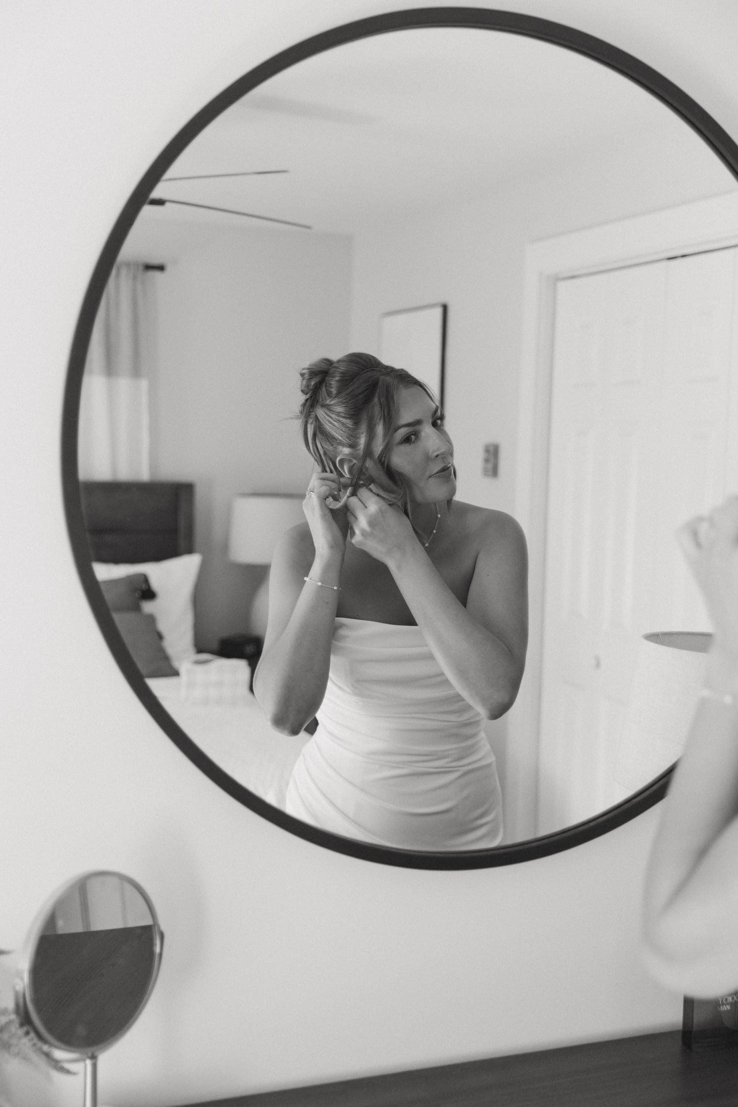 Bride adjusting her hair in a round mirror reflection, wearing a white dress in a bright room.