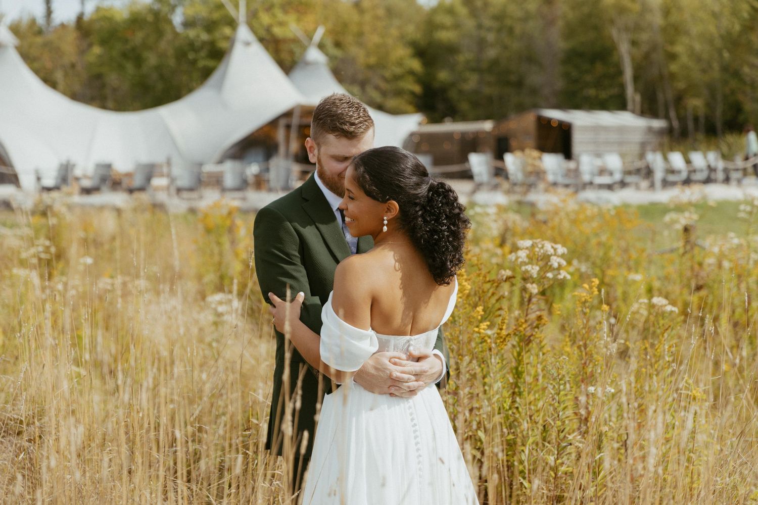 Bride and groom kissing in golden wildflower field with glamping tents visible in background.
