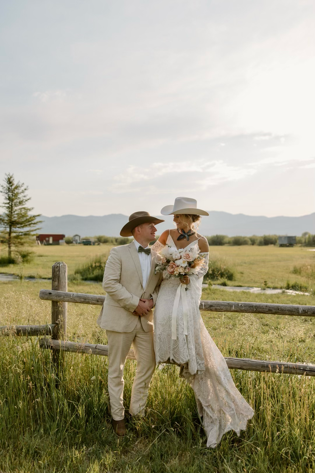 Couple in western wedding attire with cowboy hats posing by rustic fence in mountain meadow.