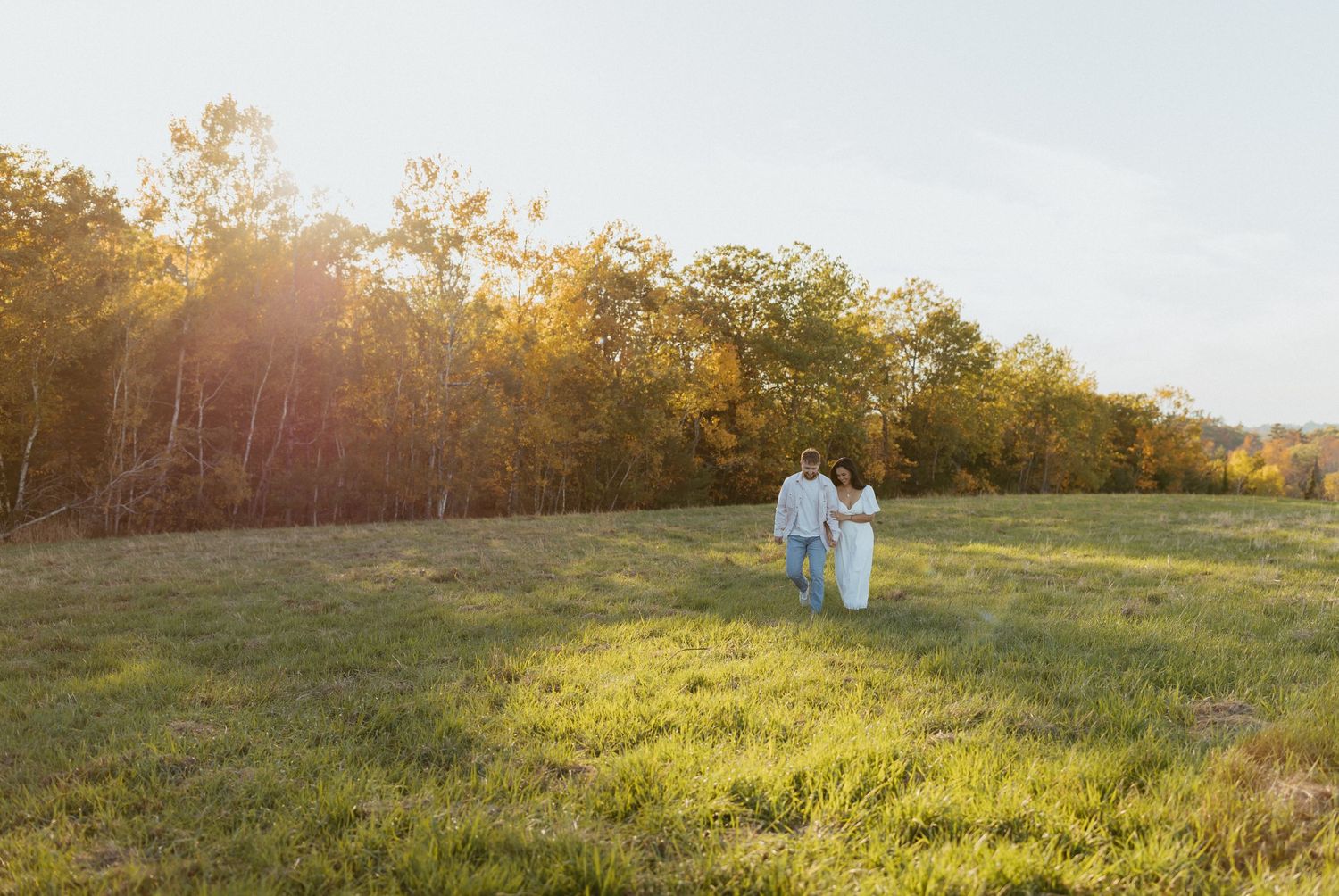 Couple walking hand in hand through a sunlit green meadow with autumn trees behind.