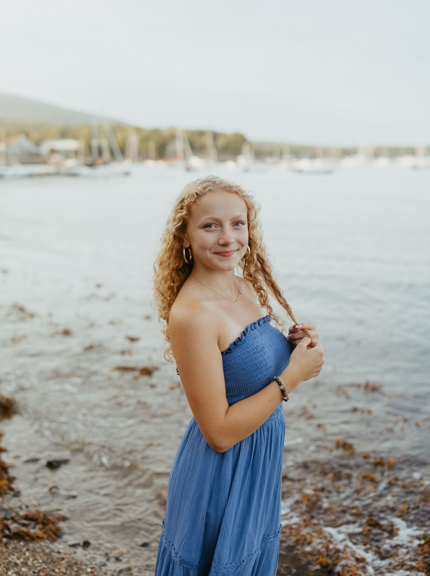 Young woman with curly hair in blue sundress smiling by the waterfront on a sunny day.