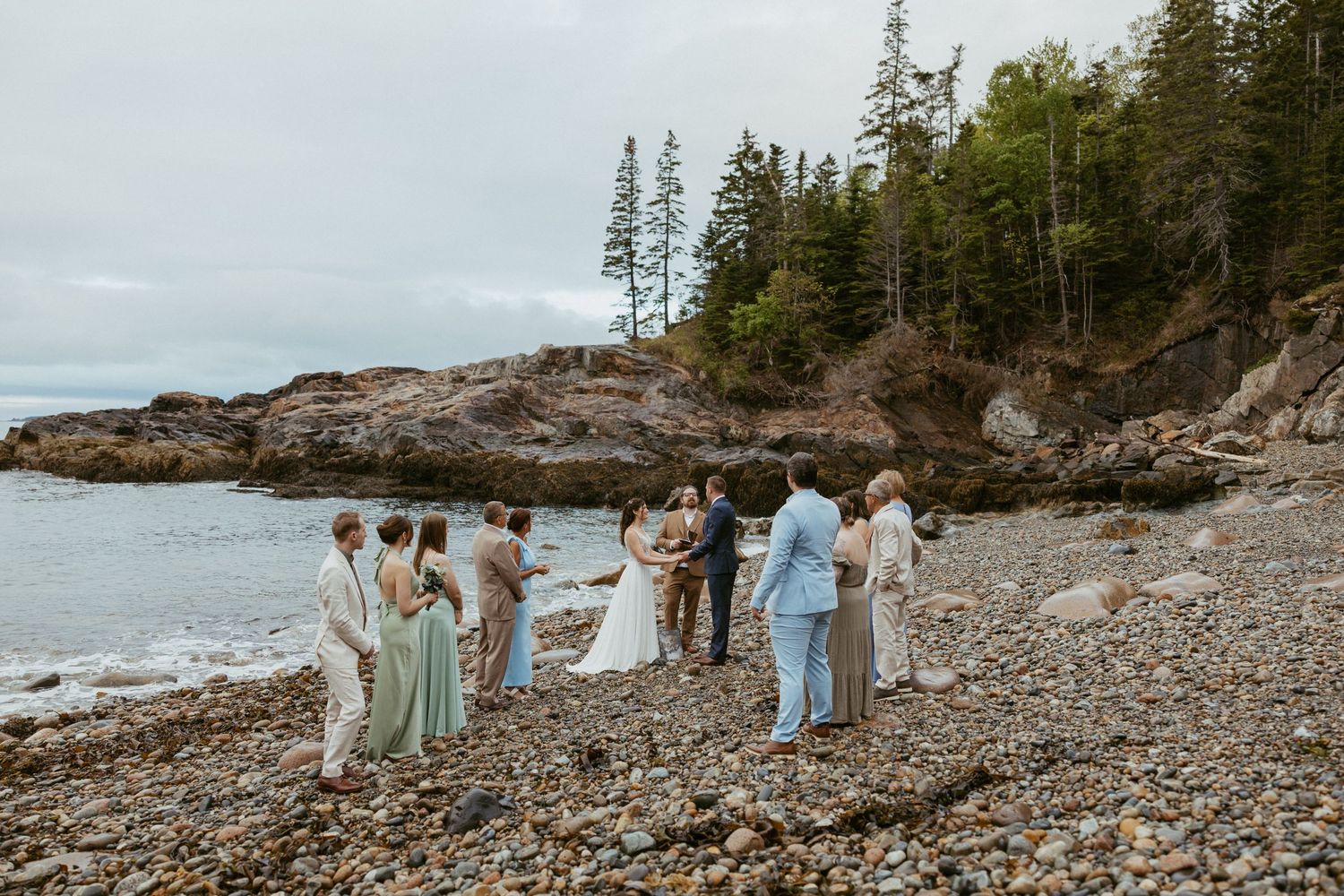 Small wedding ceremony on a rocky beach coastline with wedding party in sage green and blue attire, surrounded by ocean and pine trees.