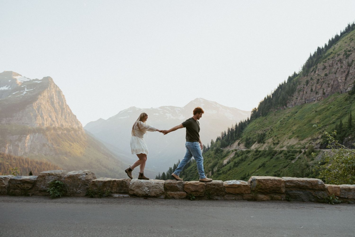 Couple holding hands walking on a stone wall with mountain scenery in the background.