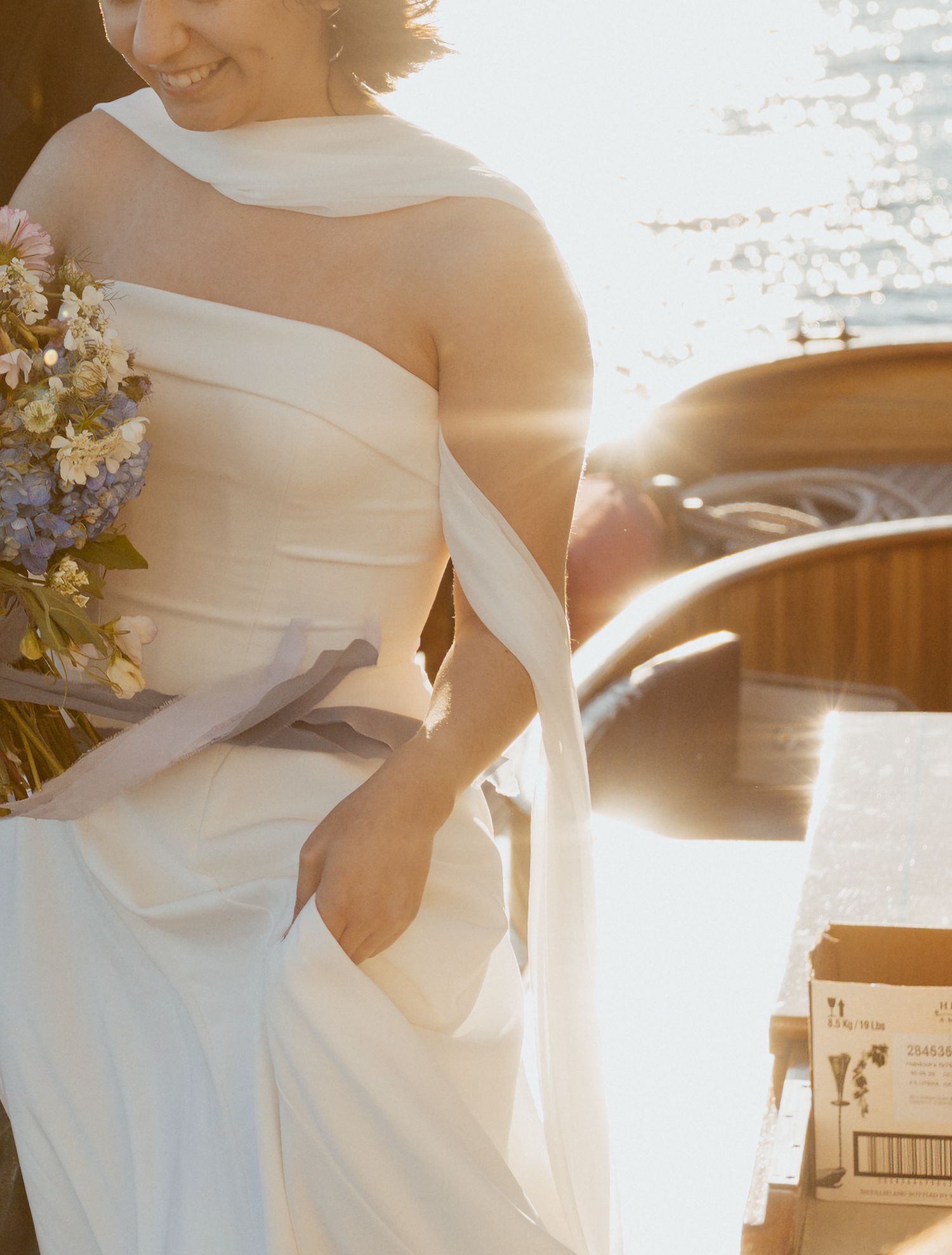 Bride in strapless white gown holding wildflower bouquet in warm sunlight on boat.