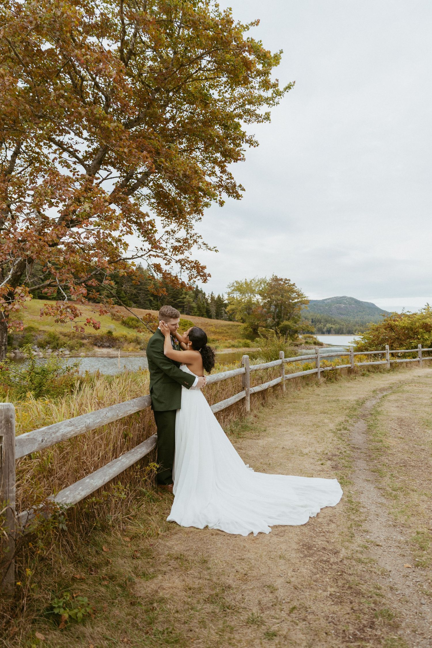 Bride and groom kissing under a colorful autumn tree beside a rustic fence near a river.