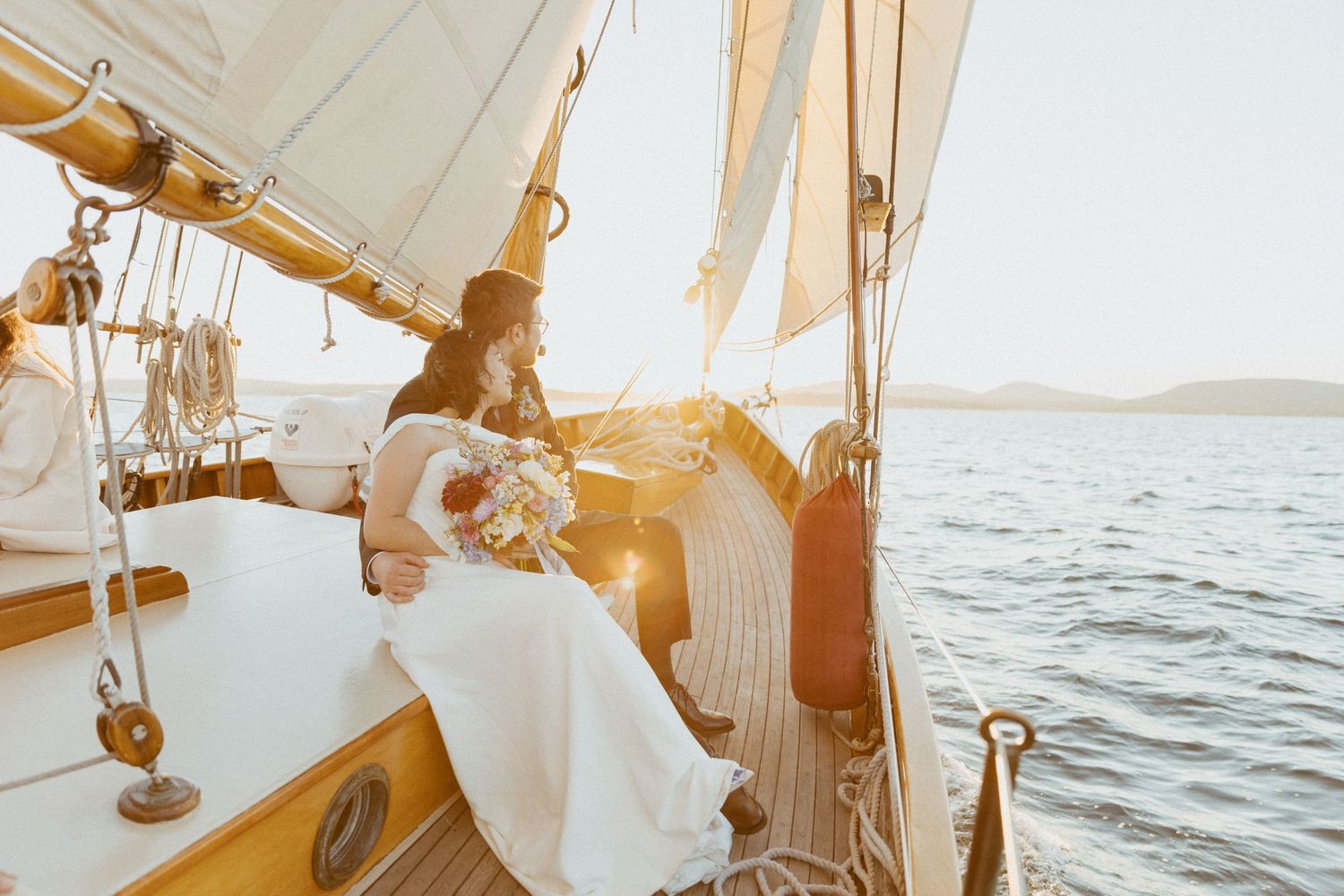 Bride in white gown holding bouquet seated on a classic wooden sailboat at golden hour sunset on calm waters.