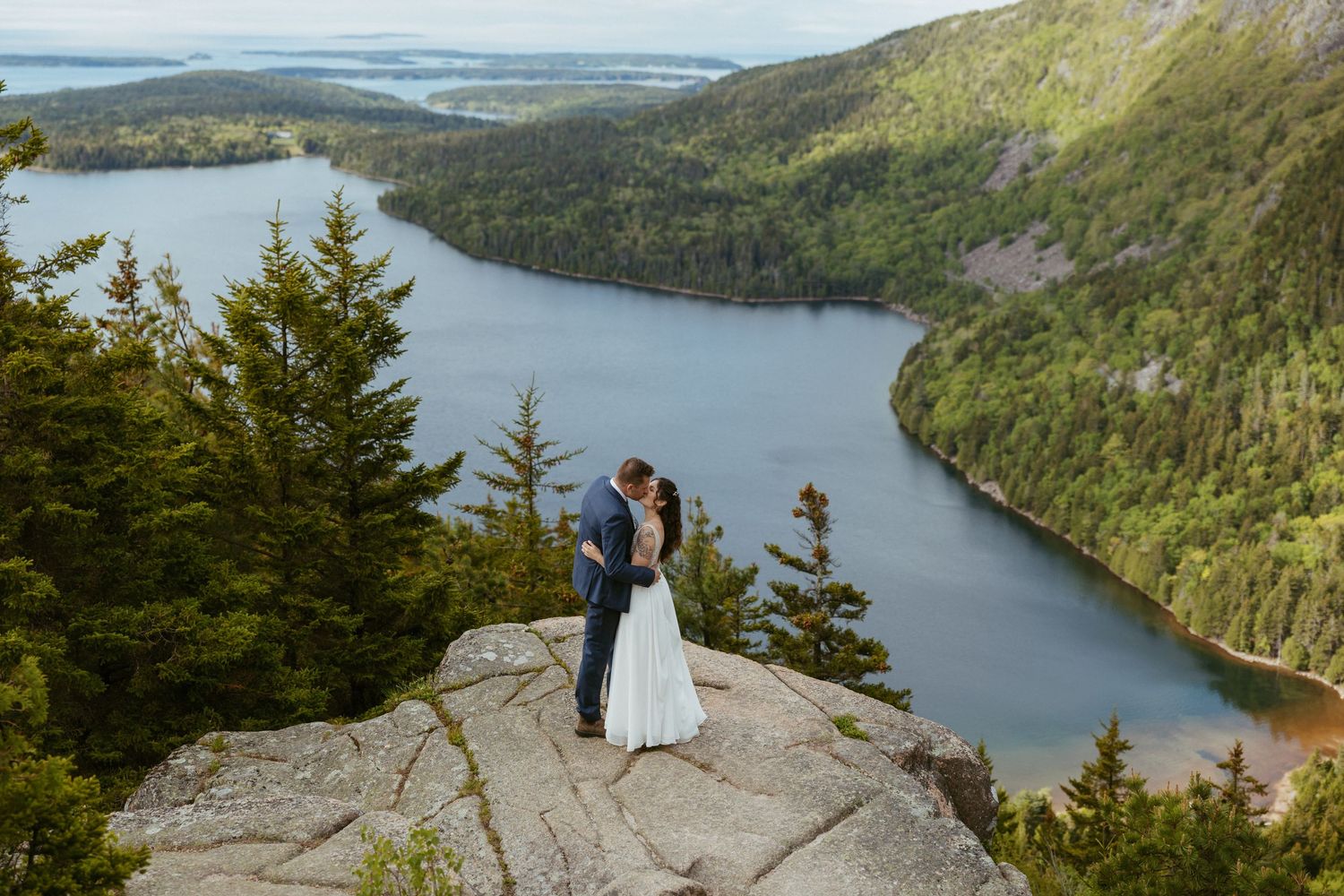 Couple shares a romantic kiss on a rocky summit overlooking a stunning lake and forested valley on their wedding day.