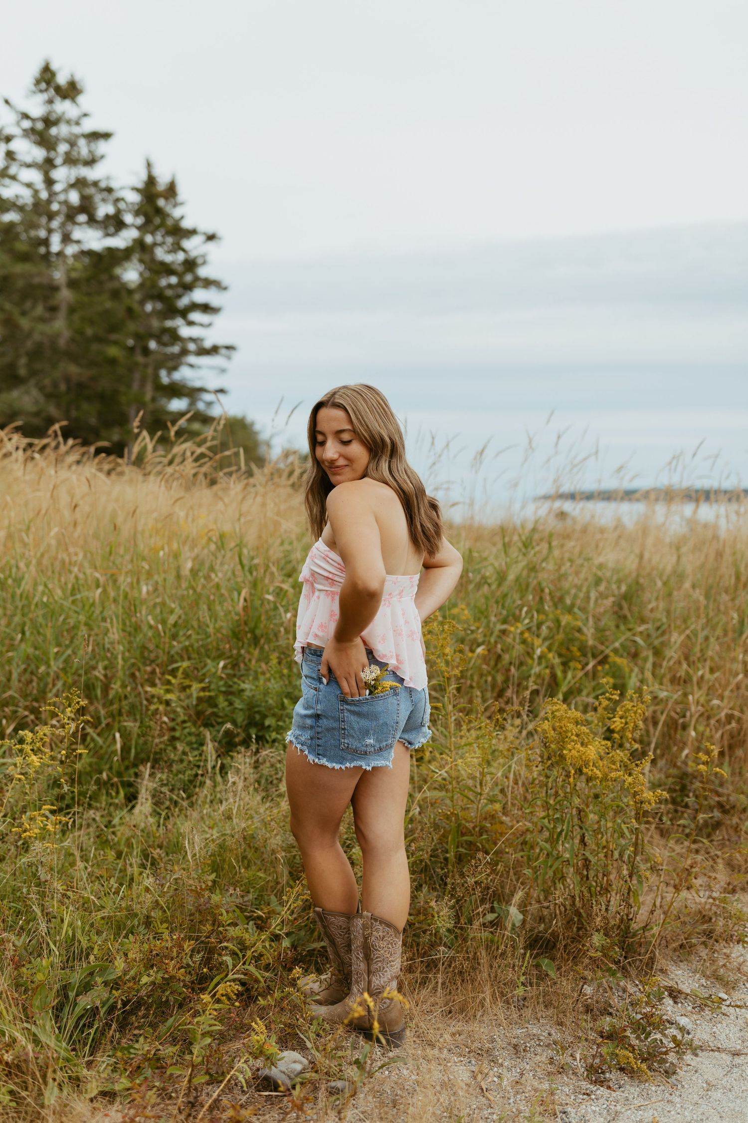 Young woman in pink top, denim shorts, and cowboy boots posing in a golden field near water.