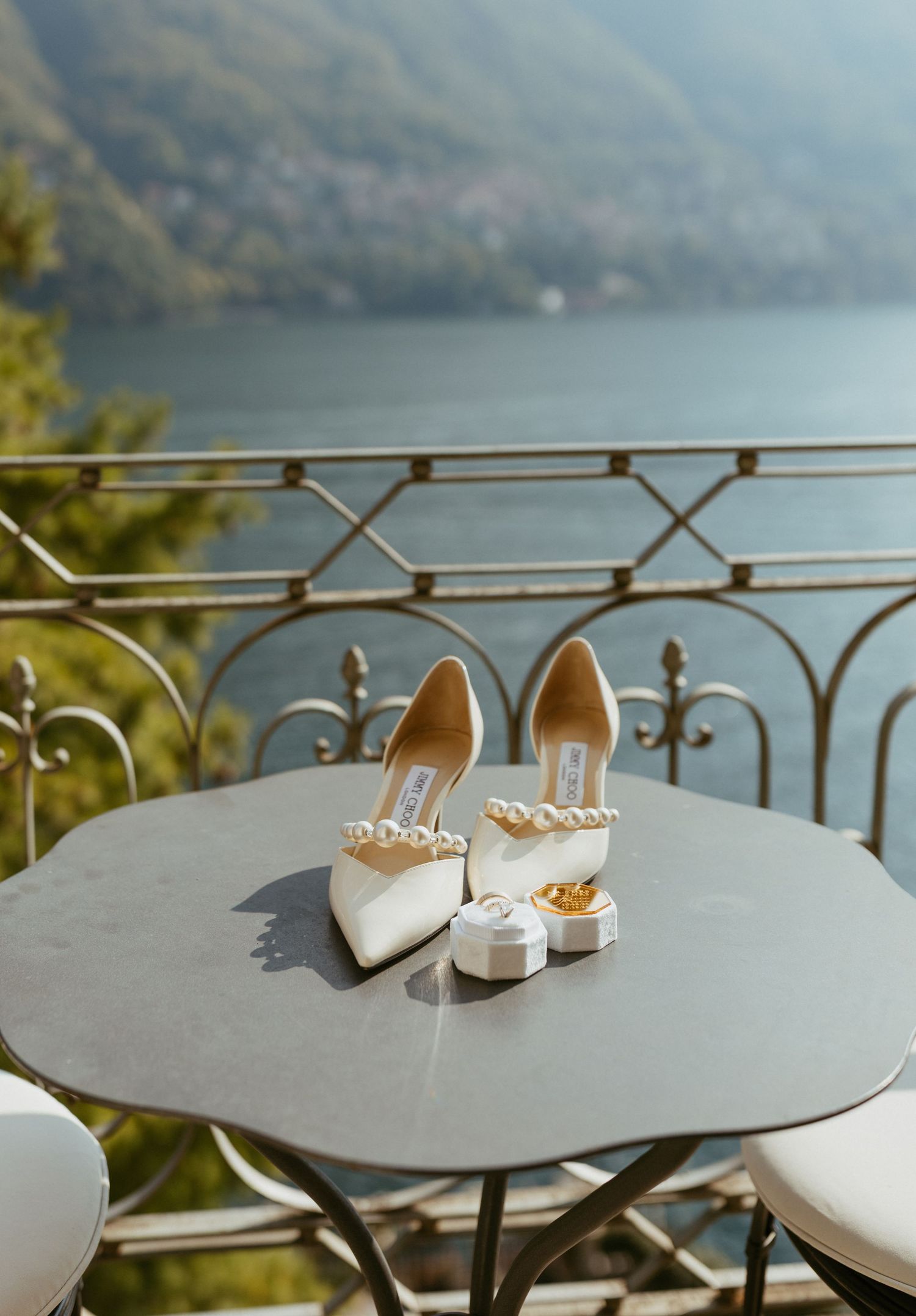 White pearl-embellished Jimmy Choo bridal heels on a balcony table overlooking a serene lake and mountains.