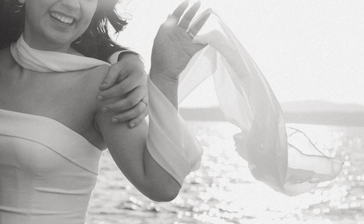 Black and white photo of a woman in a white dress on a beach, holding a flowing sheer fabric in the breeze.
