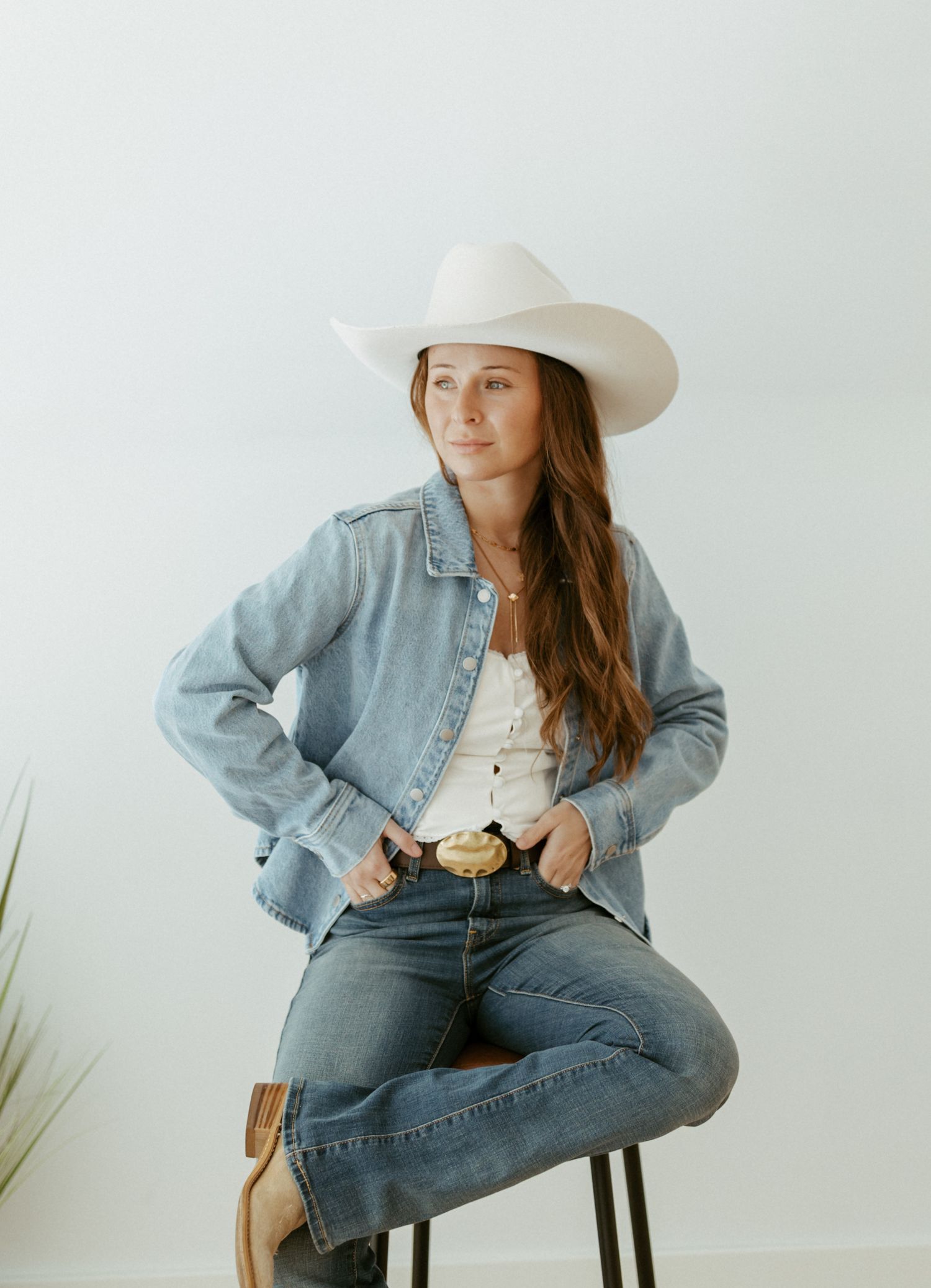 Woman in white cowboy hat and denim jacket sitting on stool wearing jeans and western boots.
