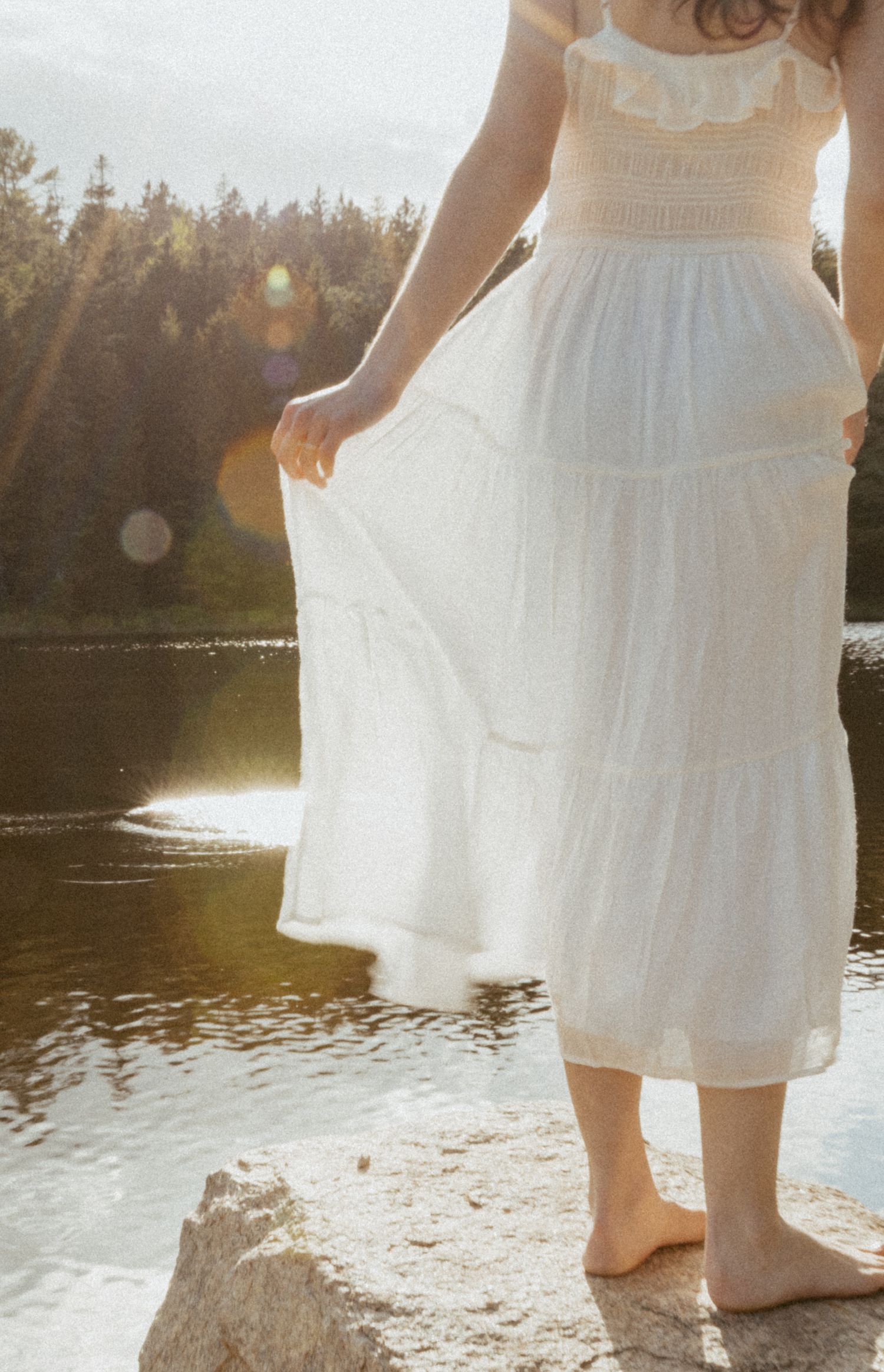 Woman in white tiered midi dress standing barefoot on rock by serene lake surrounded by forest in golden sunlight.