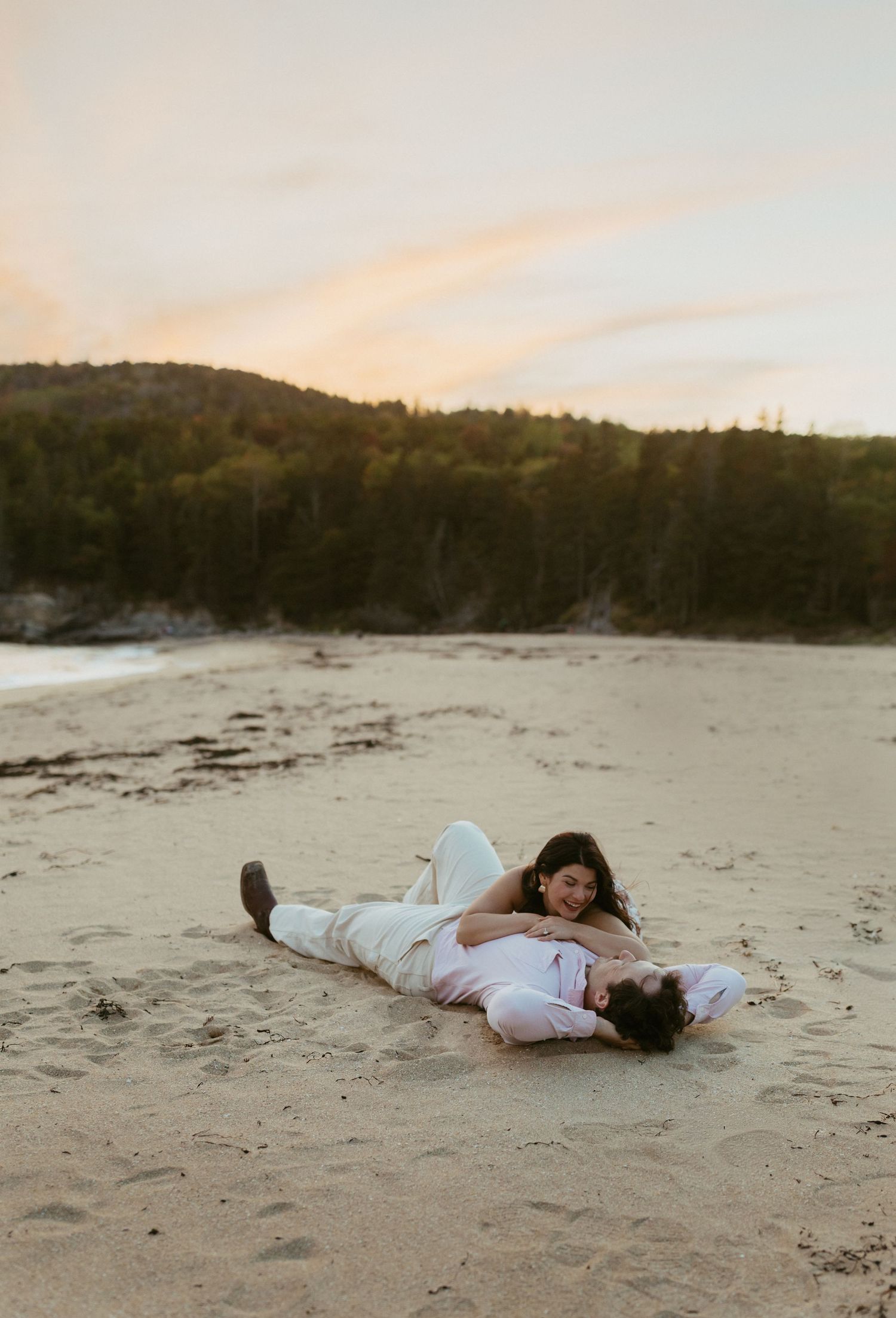 Couple lying together on Acadia sandy beach at sunset with forested hills behind them.
