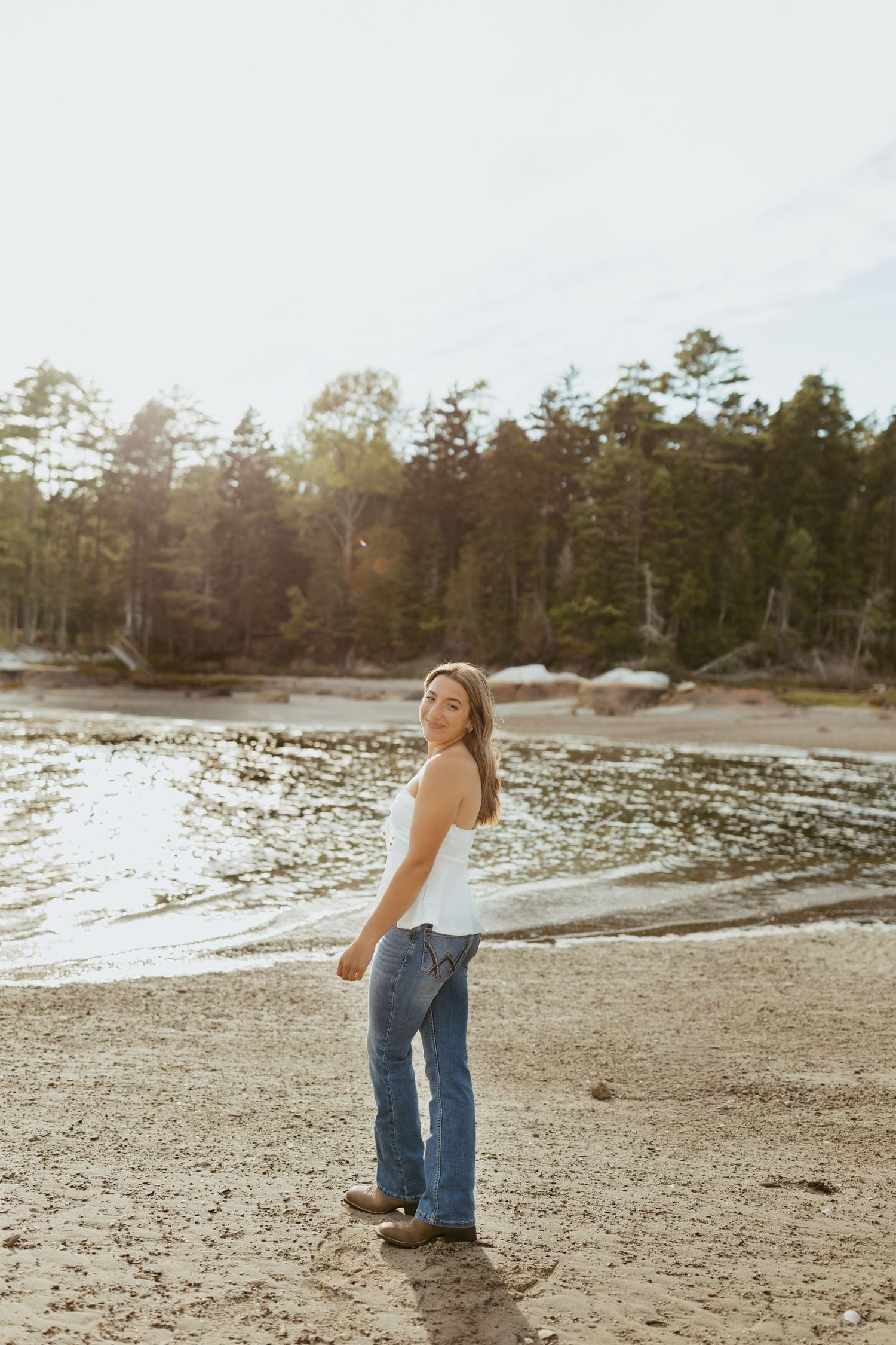 Young woman in white top and flare jeans smiling on a sandy beach with trees and water in the background.