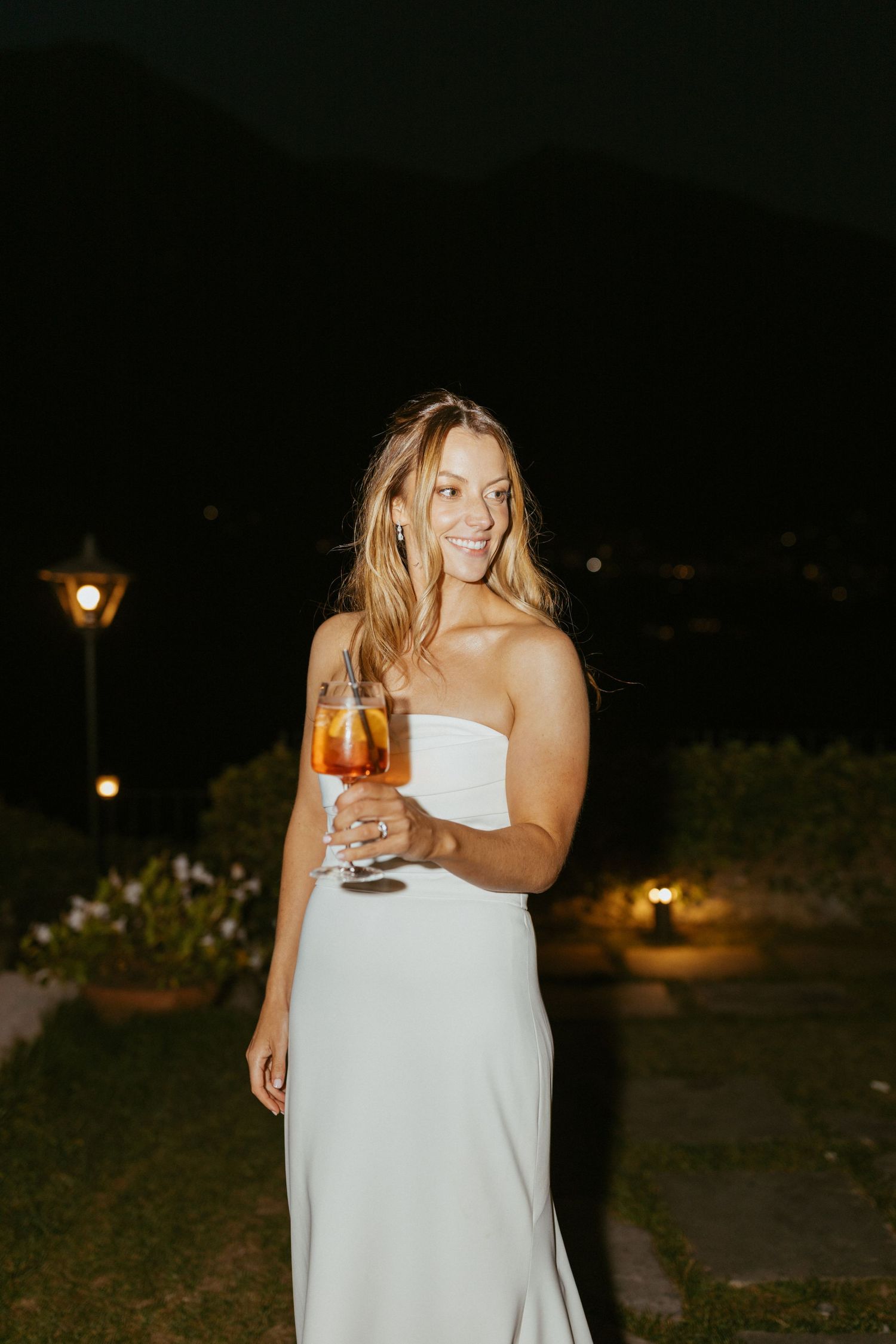 Woman in white strapless gown holding a cocktail glass outdoors at night with warm garden lights.