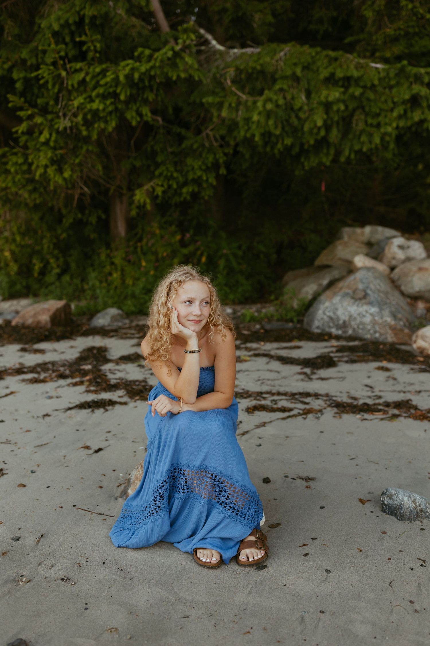 Young girl in blue maxi dress kneeling on sandy beach with rocks and green trees in background.