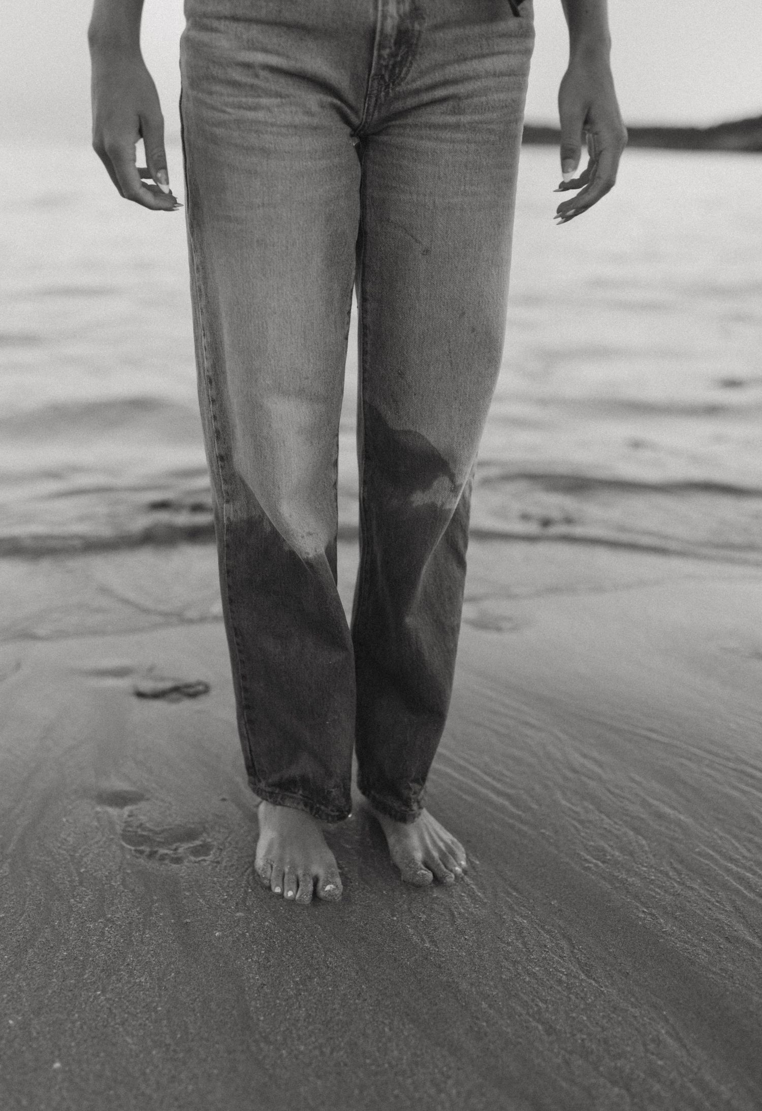 Black and white photo of bare feet and wet jeans standing on a sandy beach with ocean waves nearby.
