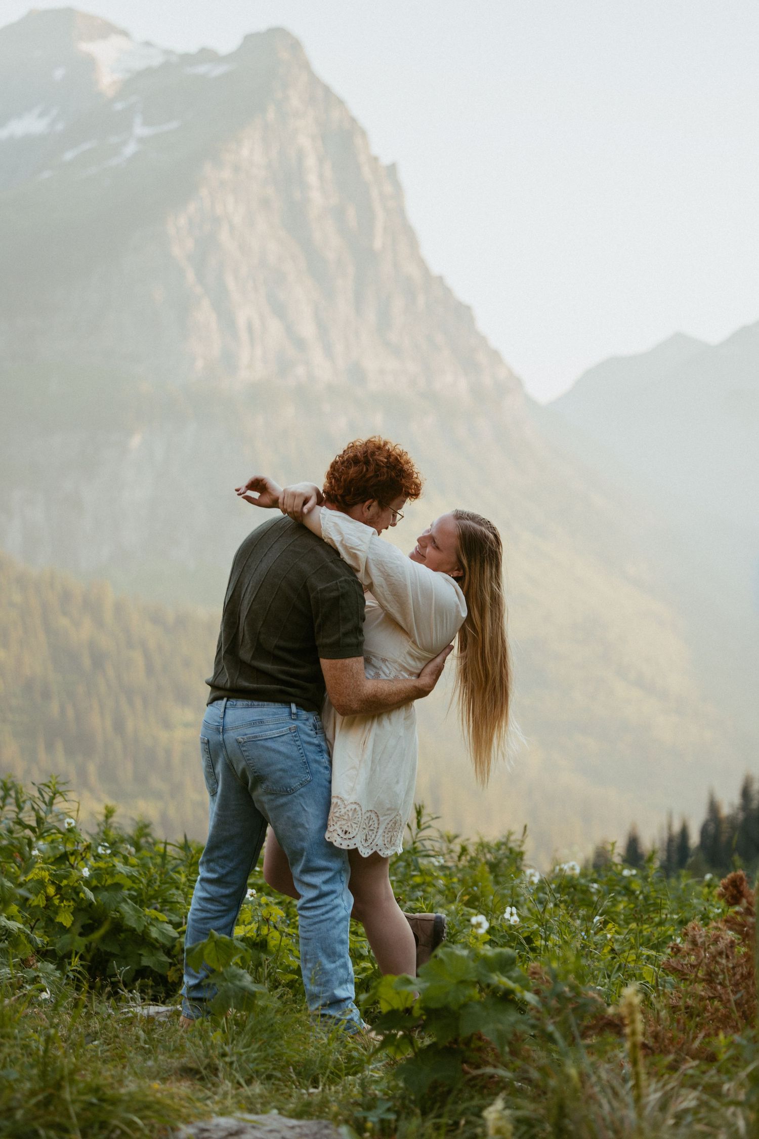 Couple dancing in a mountain meadow, woman in white dress lifted by man in jeans, scenic alpine backdrop.