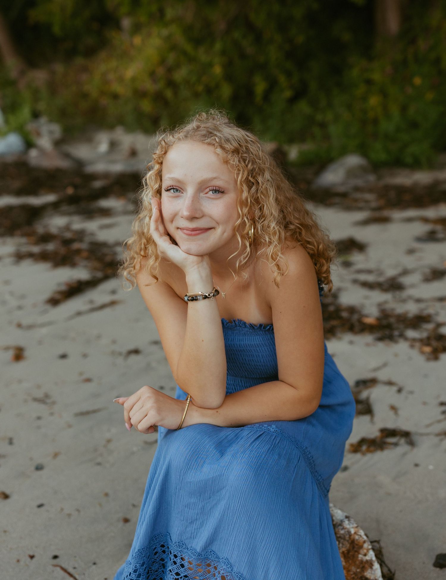 Young woman with curly blonde hair in a blue strapless dress smiling on a sandy beach.