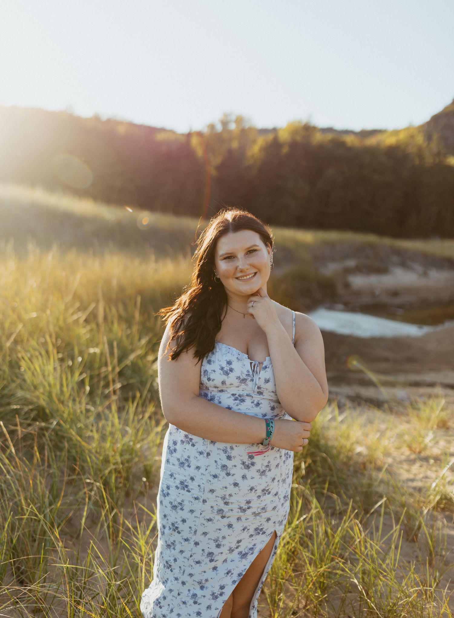 Young woman in floral sundress smiling outdoors in golden hour sunlight with grassy field and hills in background.