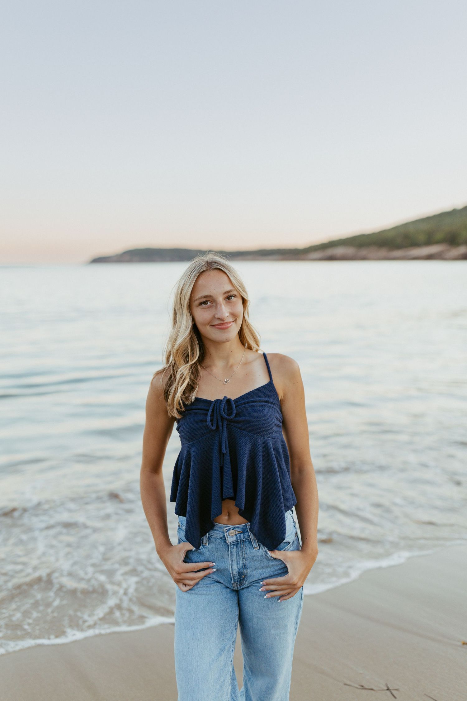 Young blonde woman in navy crop top and jeans posing on sandy beach at sunset with ocean waves behind her.