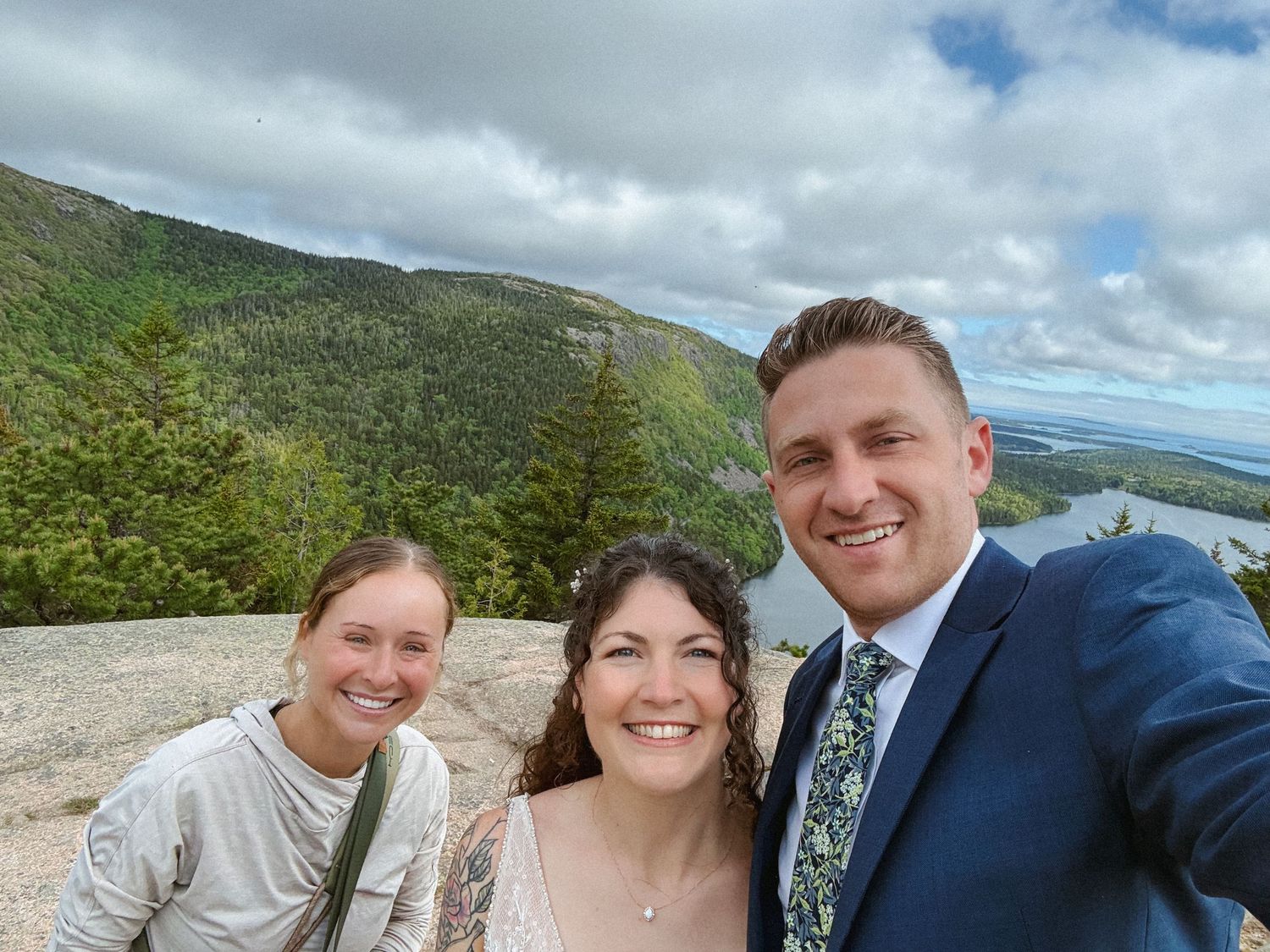 Three smiling people pose for a selfie on a mountain summit with lush green hills and cloudy sky behind them.