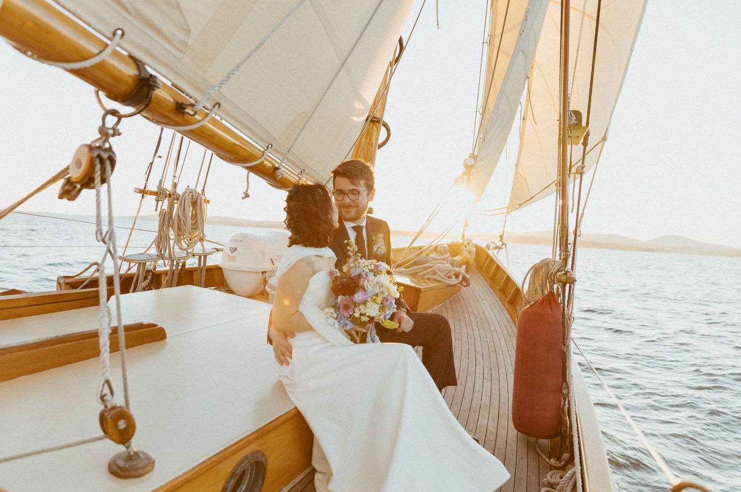 Bride and groom sharing tender moment seated on classic wooden sailboat at warm sunset.