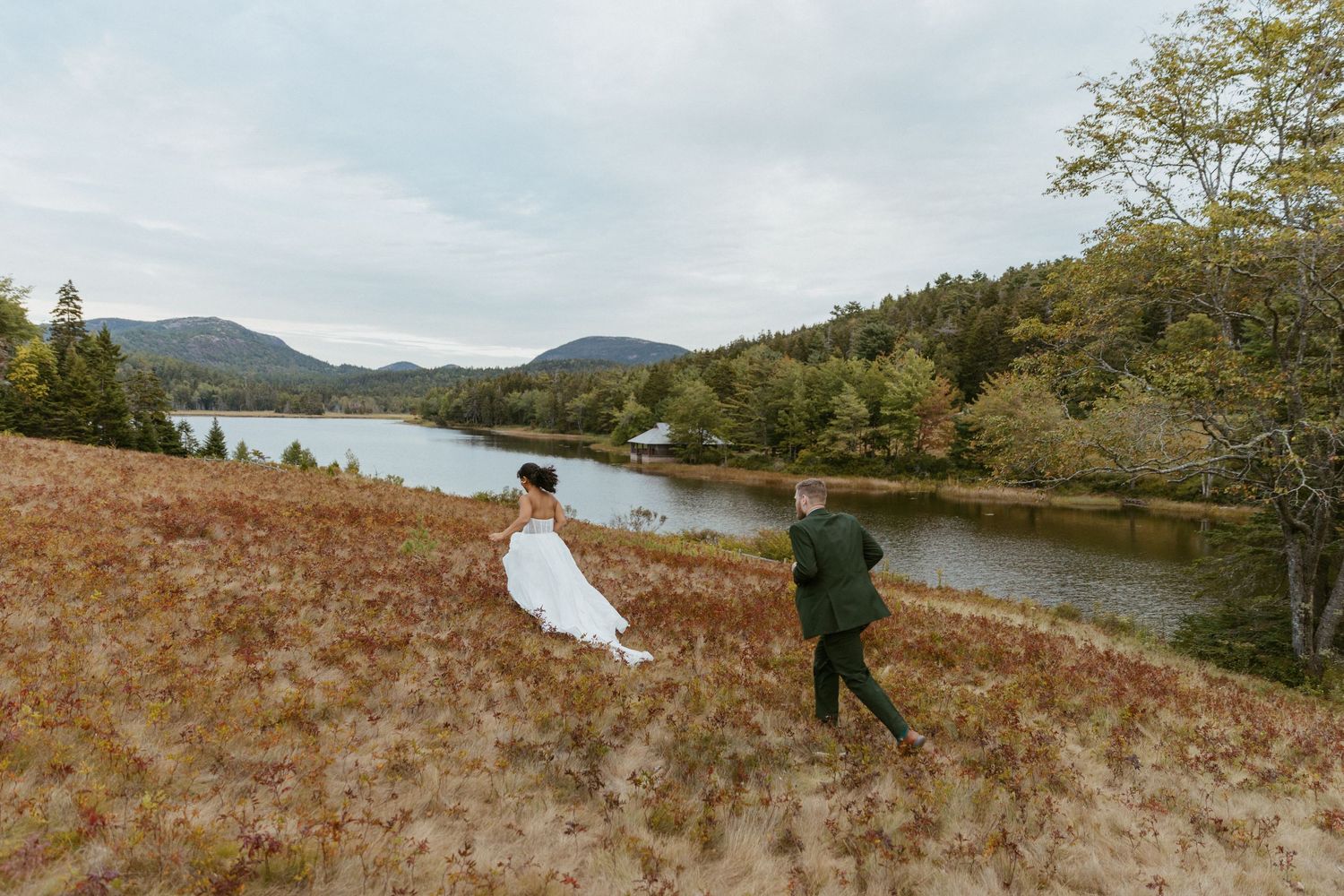 Couple in wedding attire strolling through a russet meadow beside a serene mountain lake.