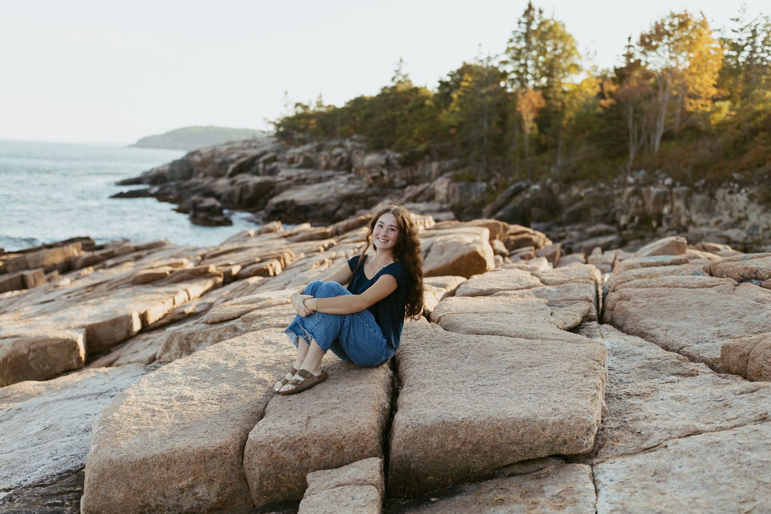 Young woman in blue dress sitting on rocky coastal shoreline with ocean and pine trees behind her.