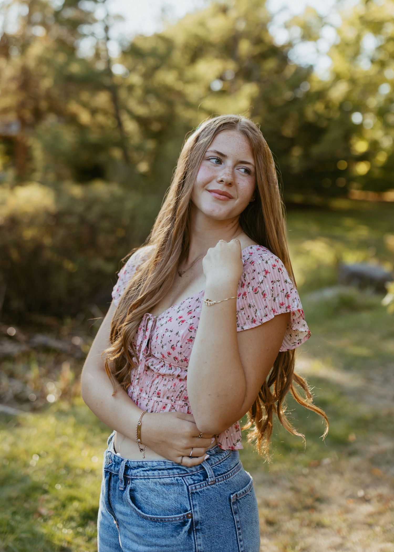 Teen girl with long auburn hair in floral crop top posing outdoors in golden sunlight.