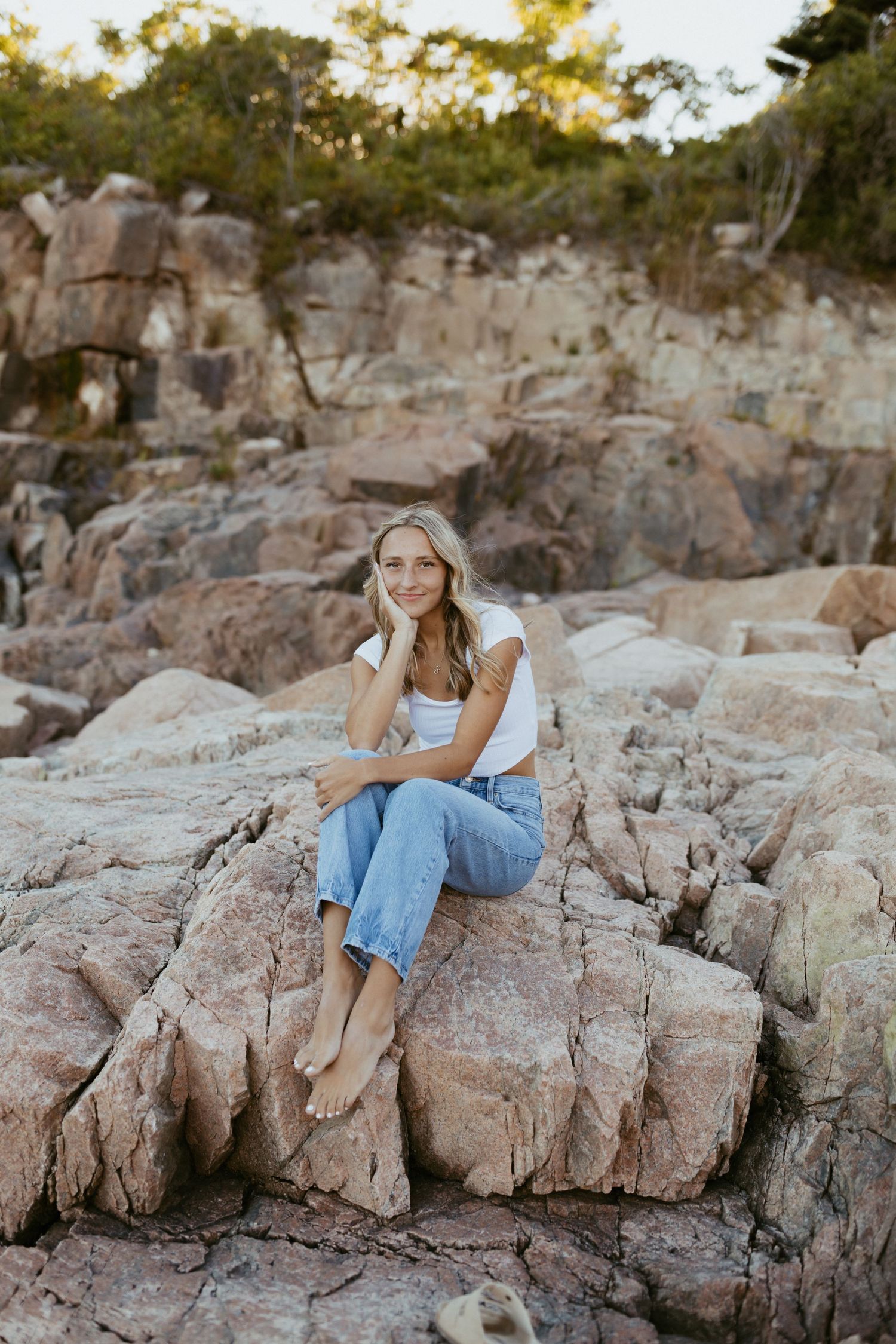 Young blonde woman sitting barefoot on rocky coastal terrain, wearing white top and blue jeans, smiling at camera.