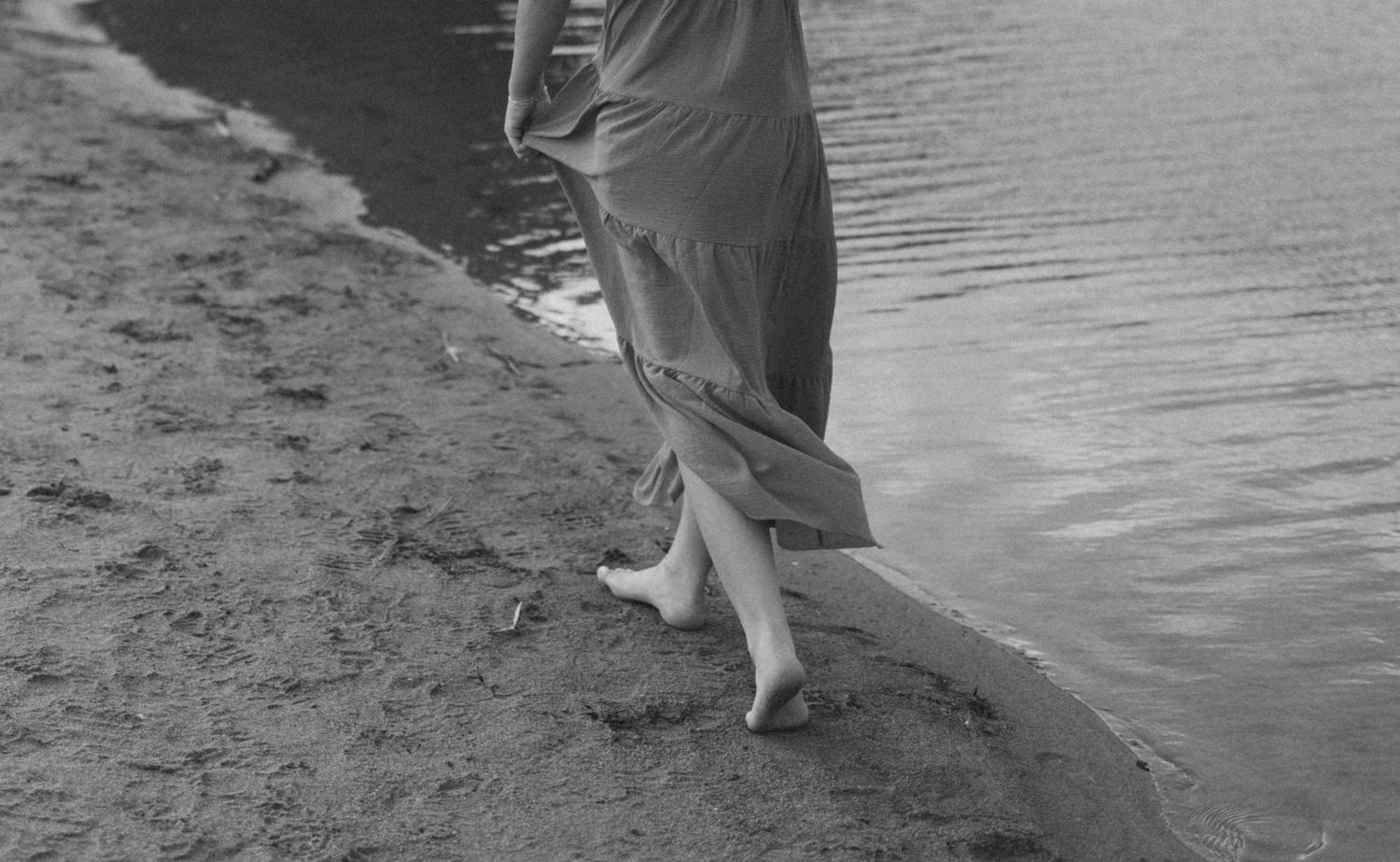 Black and white photo of a woman in a long dress walking barefoot along a sandy shoreline near calm water.