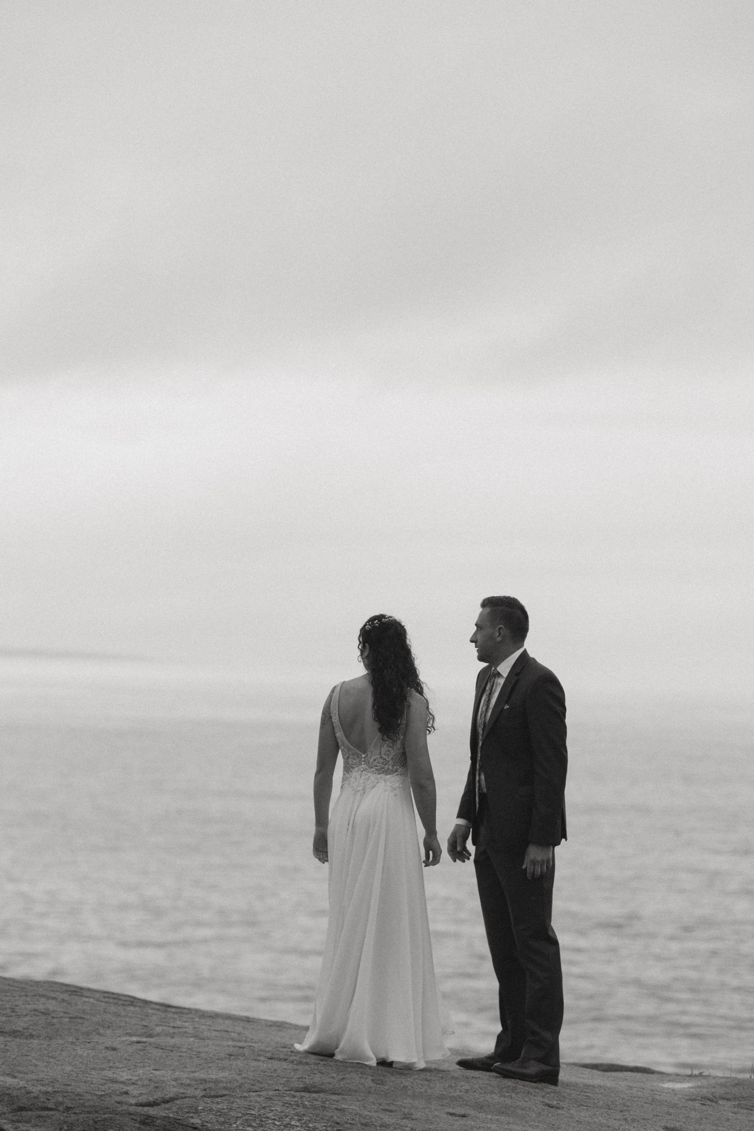 Black and white photo of a bride and groom standing on a cliff overlooking the ocean on their wedding day.