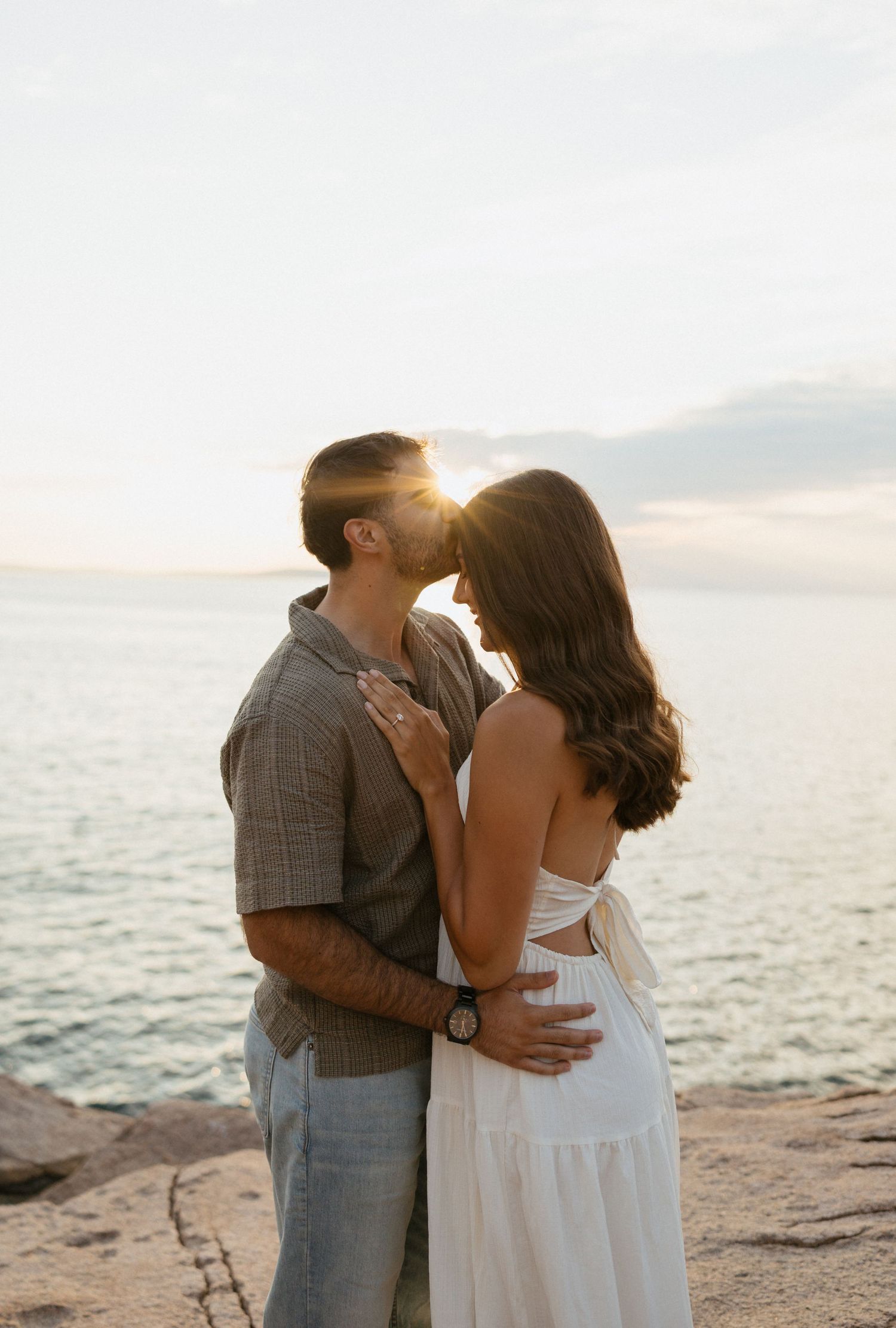 Couple sharing a romantic sunset kiss by the ocean, woman in white dress, golden hour light glowing behind them.