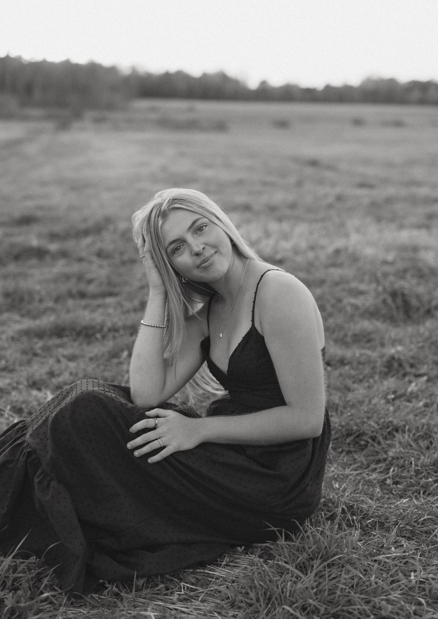 Black and white photo of a young blonde woman in a dark dress sitting gracefully in an open grassy field.