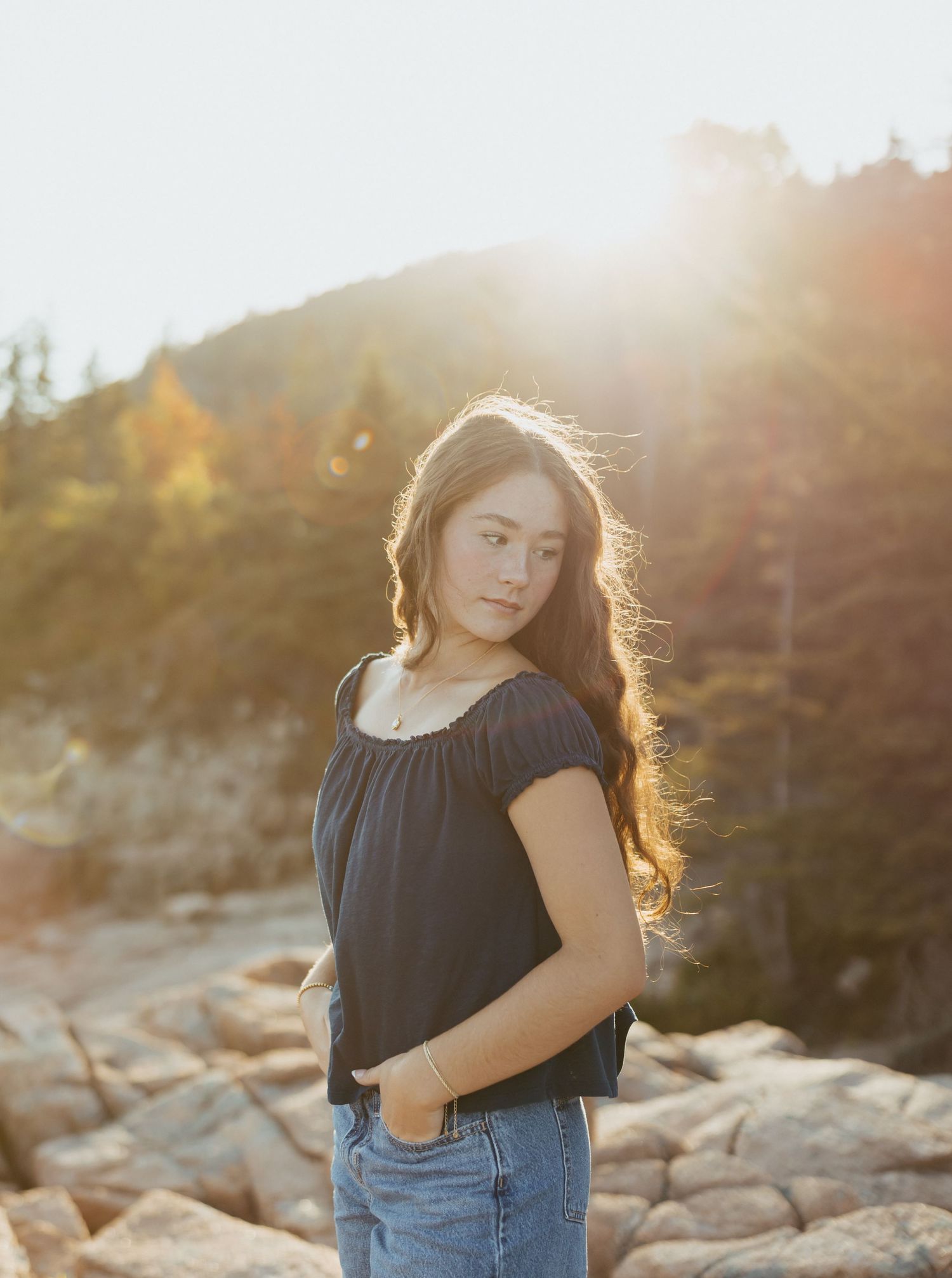 Young woman in dark top and jeans poses on rocky terrain with golden sunlight and trees in background.