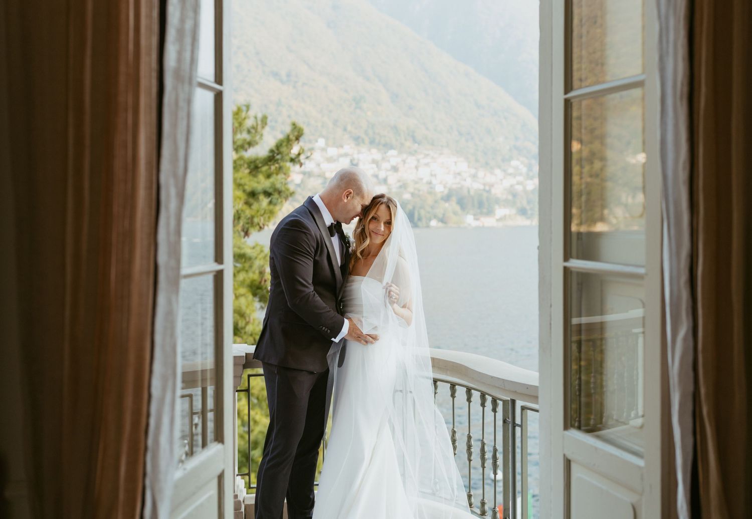 Bride and groom share romantic kiss on balcony overlooking scenic lake and mountains on wedding day.