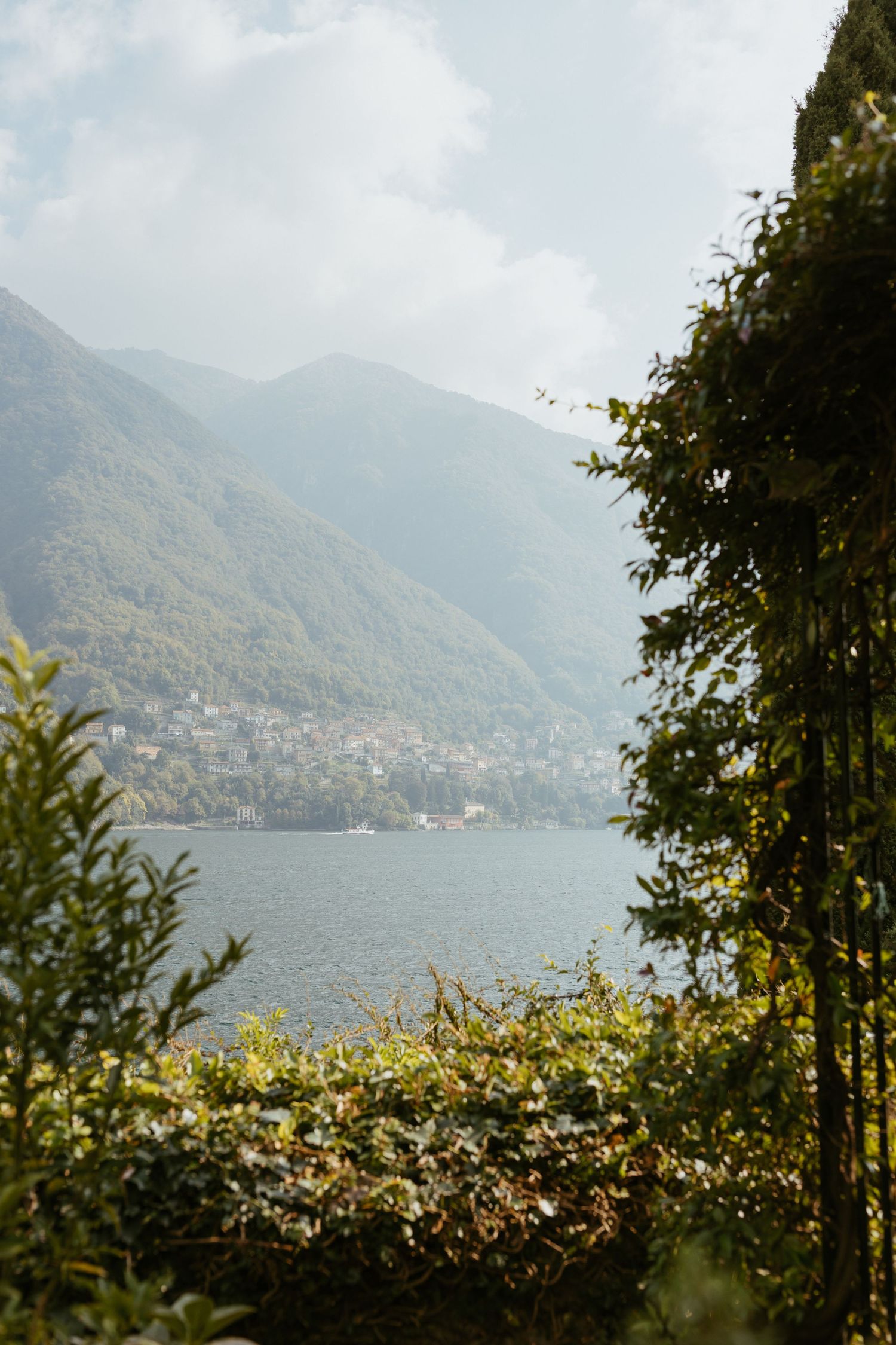 Scenic view of Lake Como framed by lush green trees with misty mountains and a lakeside village in the background.