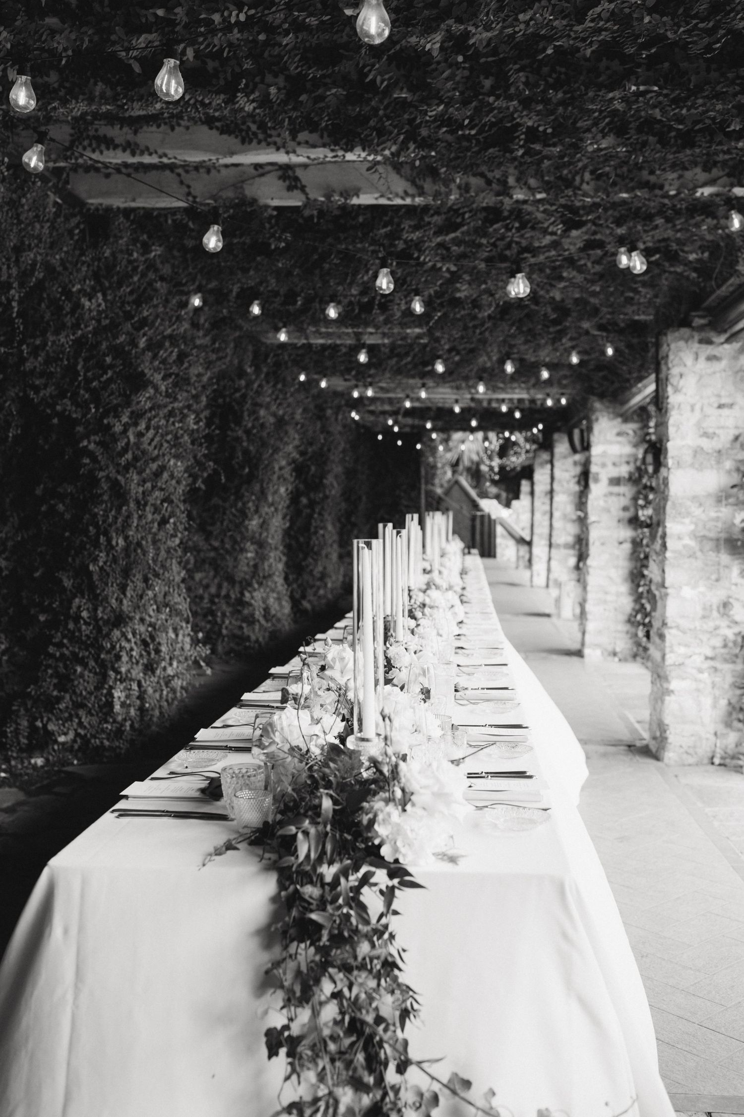Black and white photo of an elegant long wedding reception table with floral garland, candles, and string lights under ivy pergola.