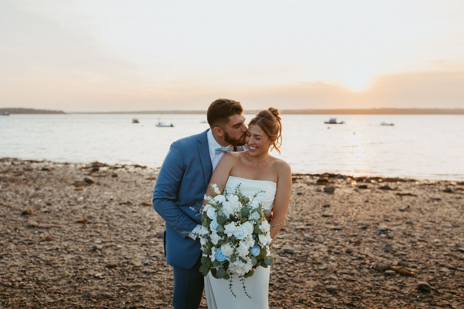 Bride and groom share a romantic sunset kiss on a rocky Maine beach, bride holding a white and blue floral bouquet.