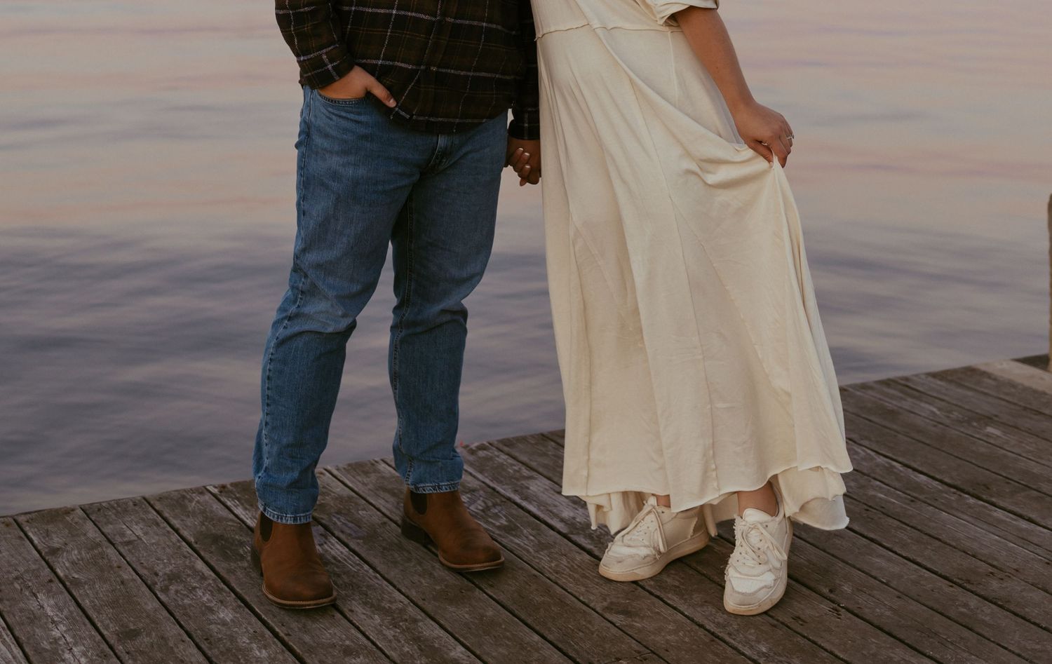 Close-up of couple holding hands on a wooden dock, woman in white dress and man in jeans.