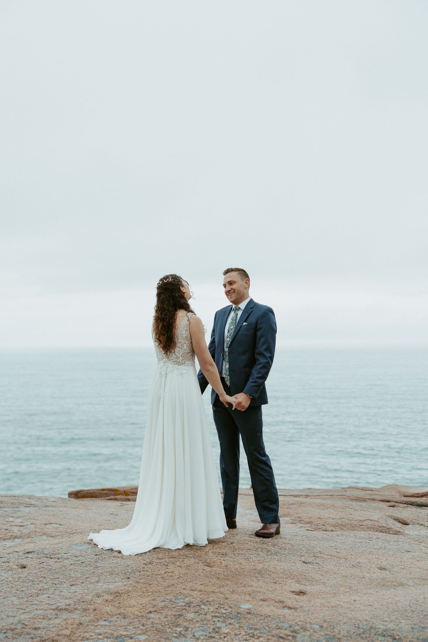 Bride and groom hold hands on rocky ocean cliff, groom in navy suit smiling at bride in white gown.